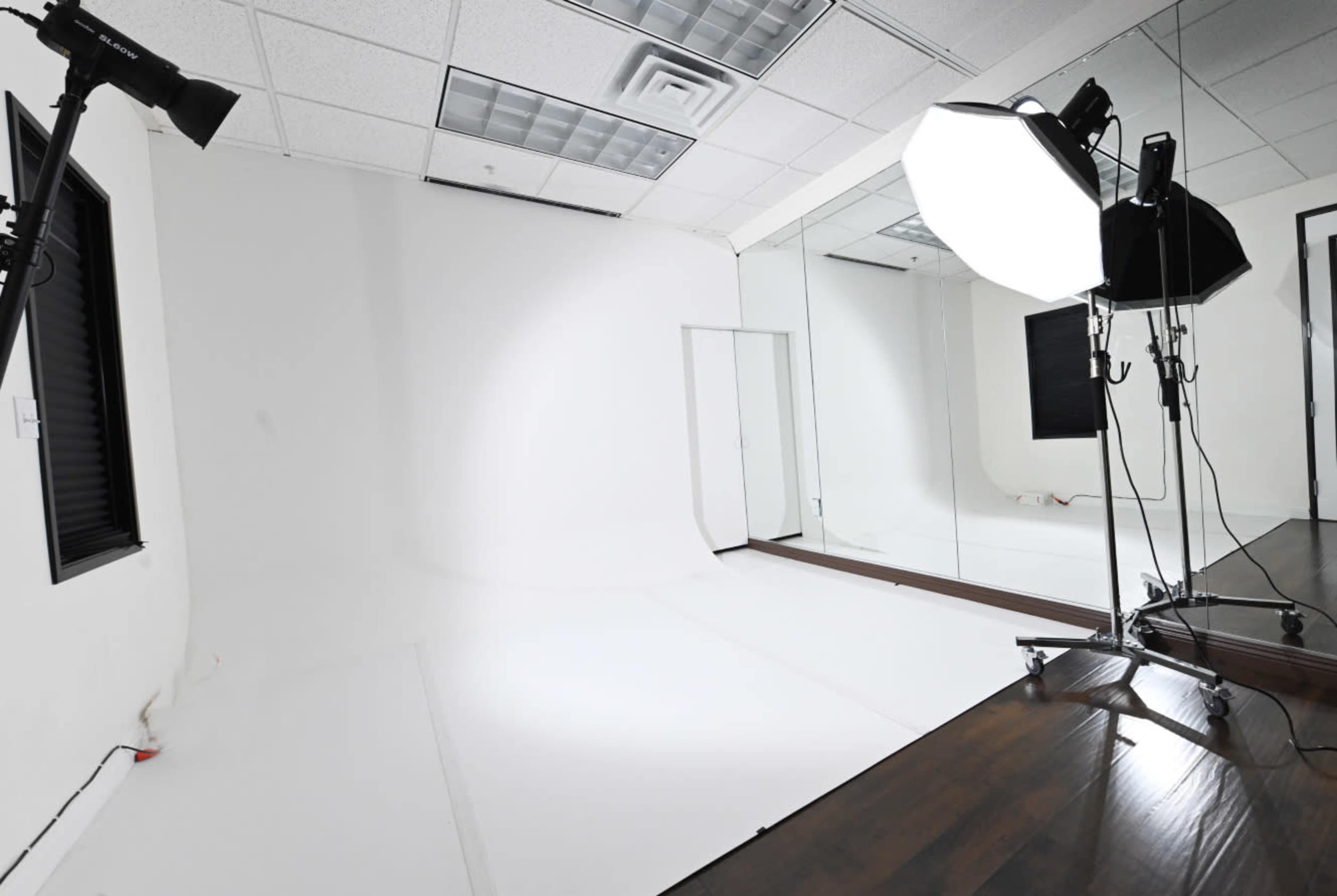 The image shows a photography studio with a white background, a large softbox light, and a mirrored wall.