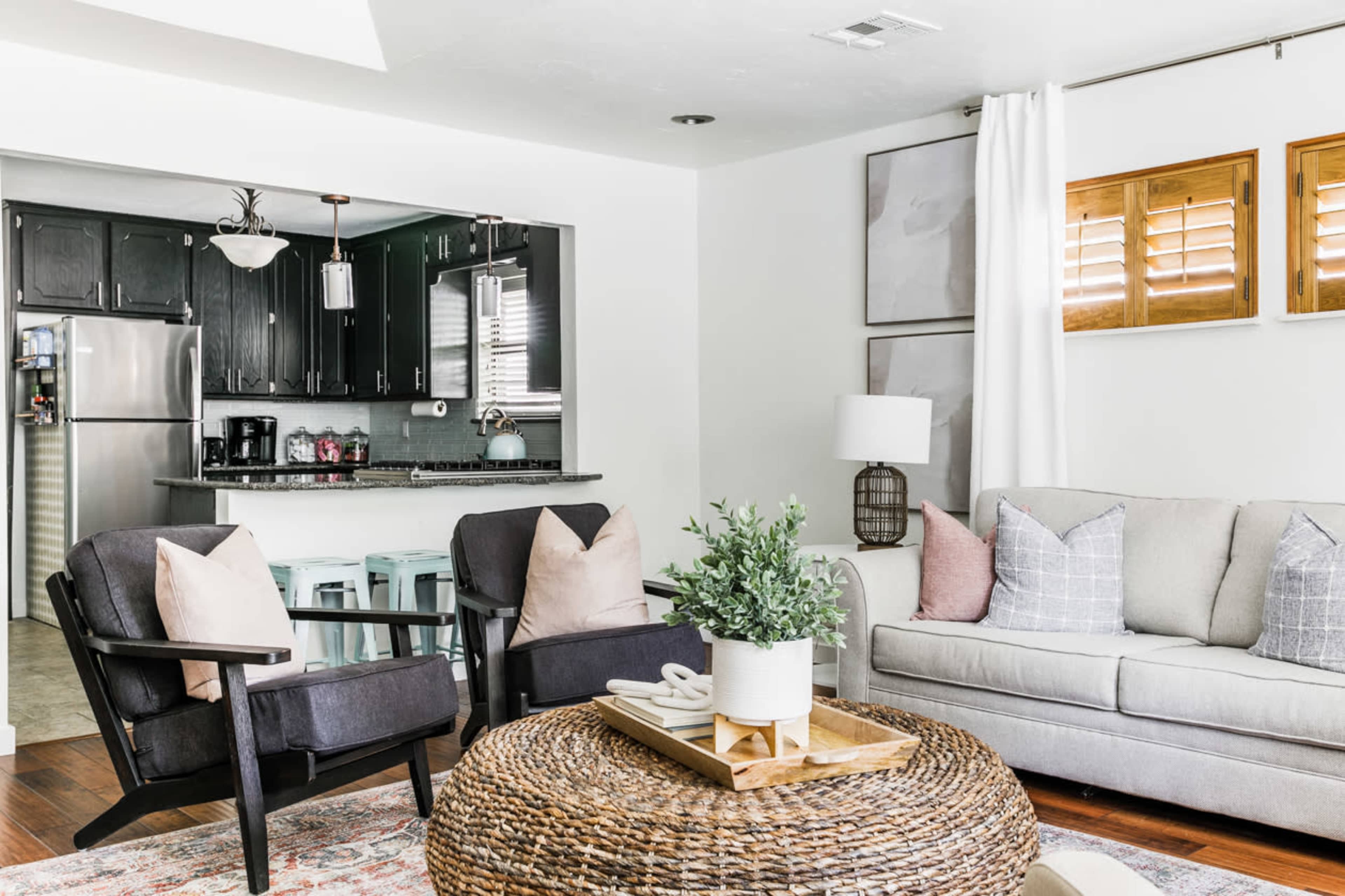 A modern living room with a gray sofa, two black armchairs, a round woven coffee table, and a kitchen area visible in the background.
