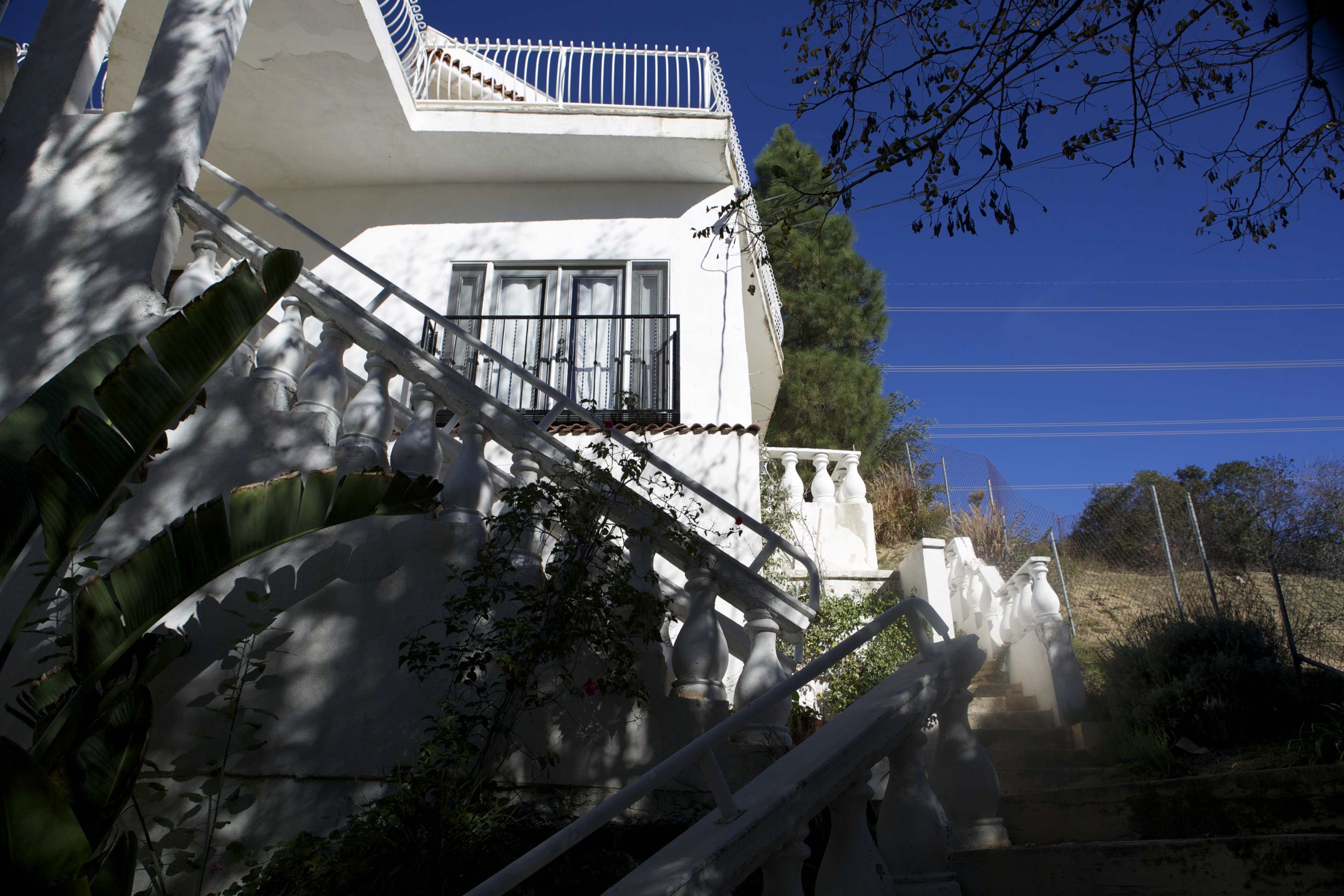 A white house with a balcony is situated on a hillside, featuring a staircase bordered by greenery.