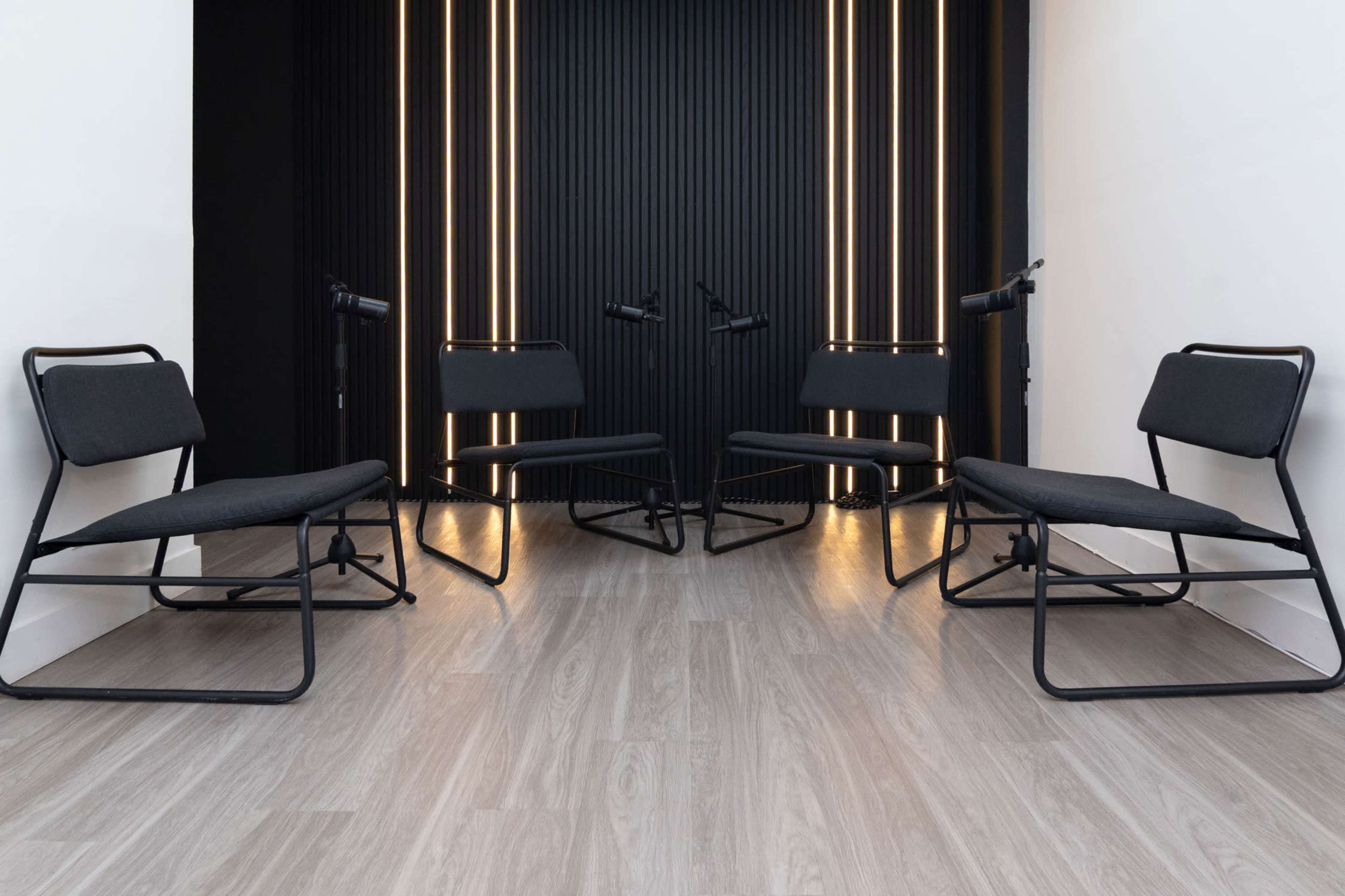 The image shows a small, modern meeting space with four black chairs arranged in a circle, against a backdrop of vertical black panels with backlighting.