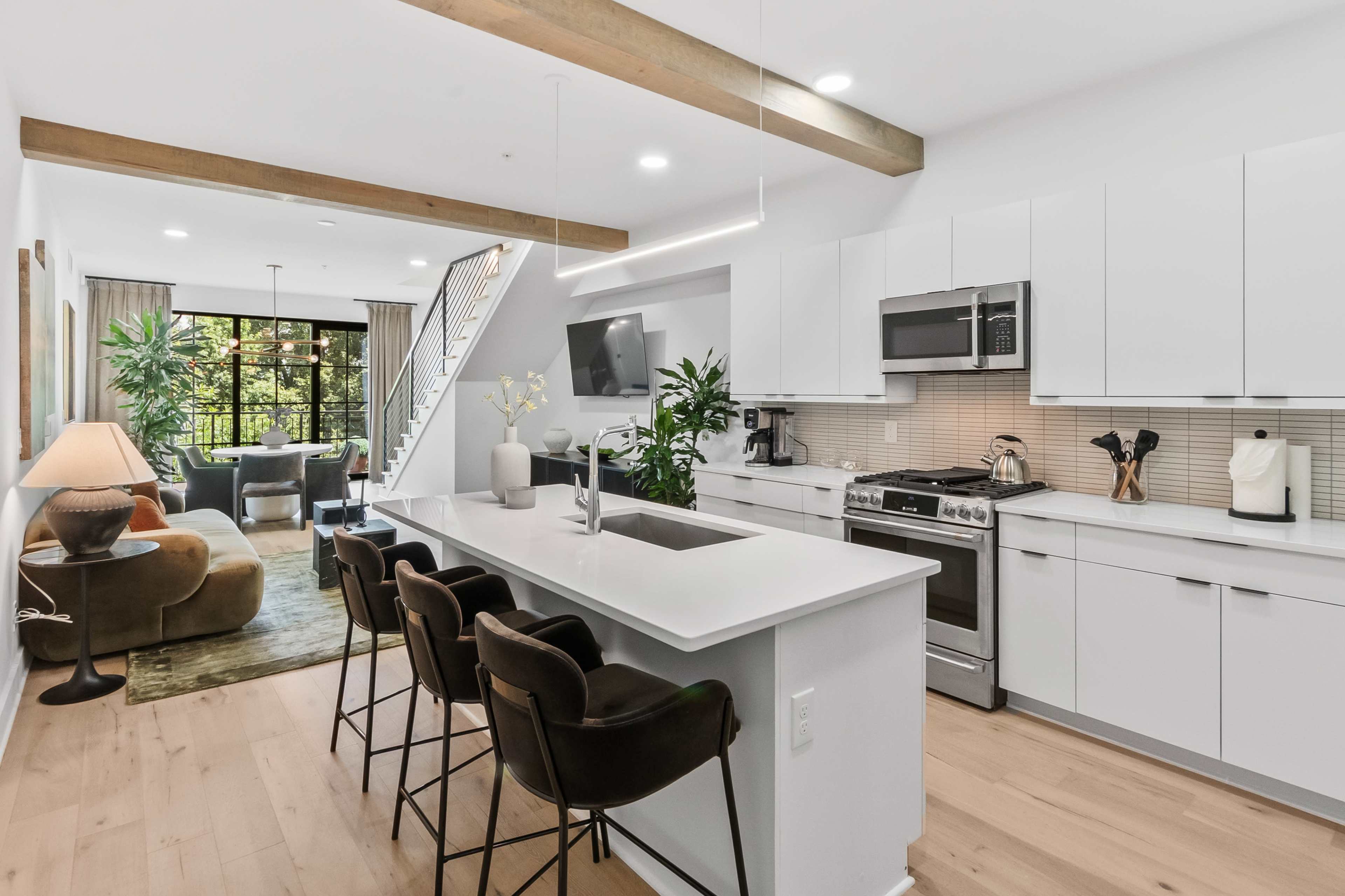The image shows a modern kitchen with white cabinetry, a central island with bar stools, and large windows that provide natural light to the adjacent living area.