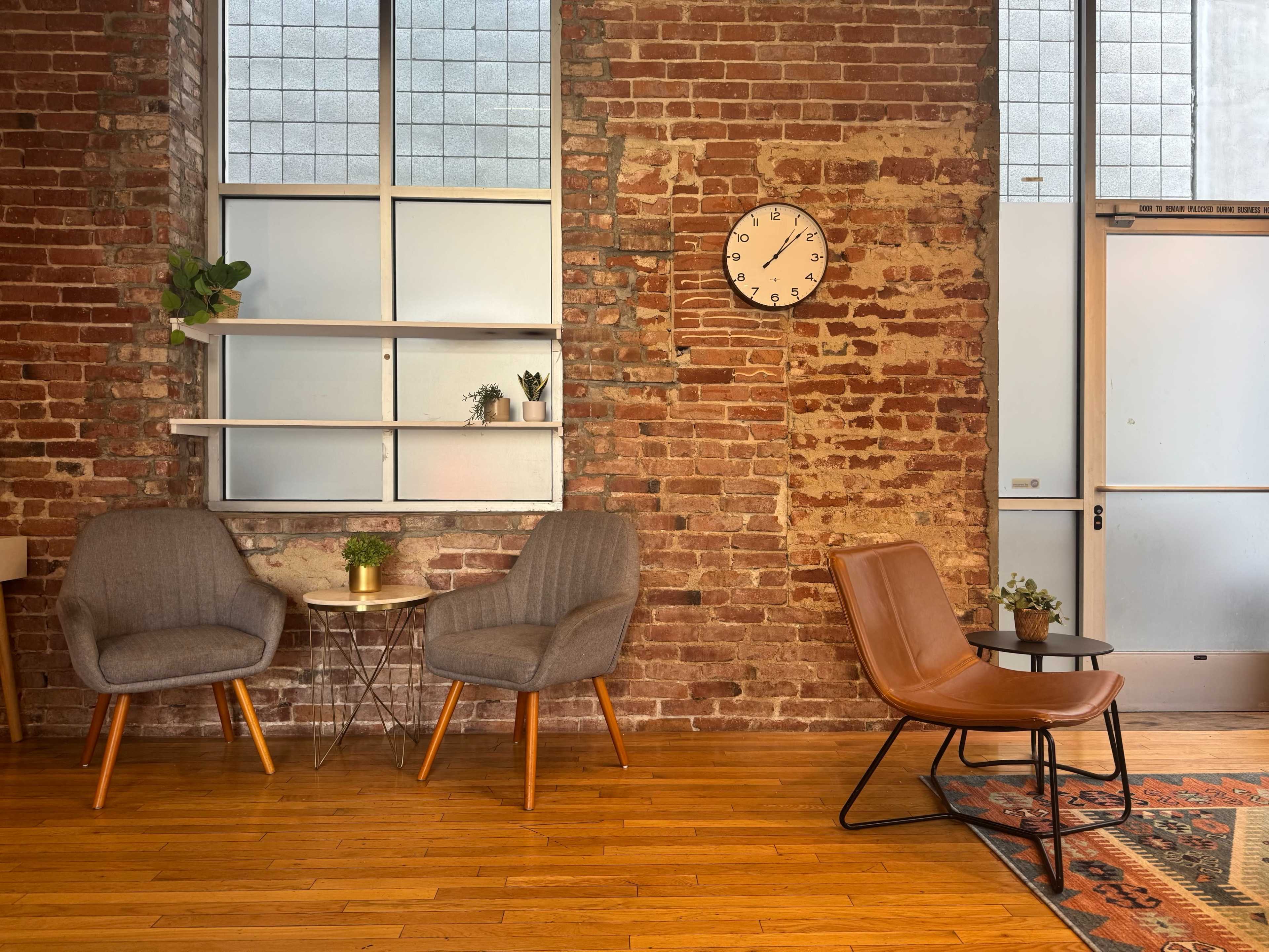 The image shows a cozy space featuring two chairs, a small table, a wall clock, and exposed brick walls.