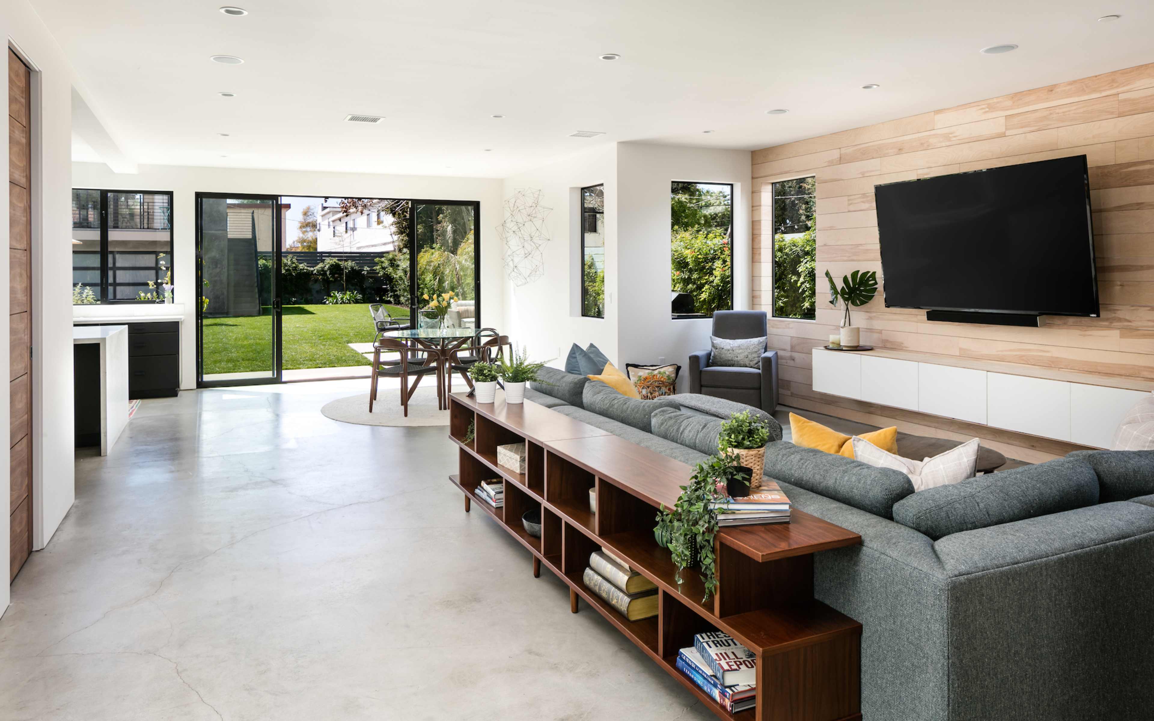 The image shows a modern living room with a large sofa, a wooden shelf, and a view of a grassy backyard through expansive glass doors.