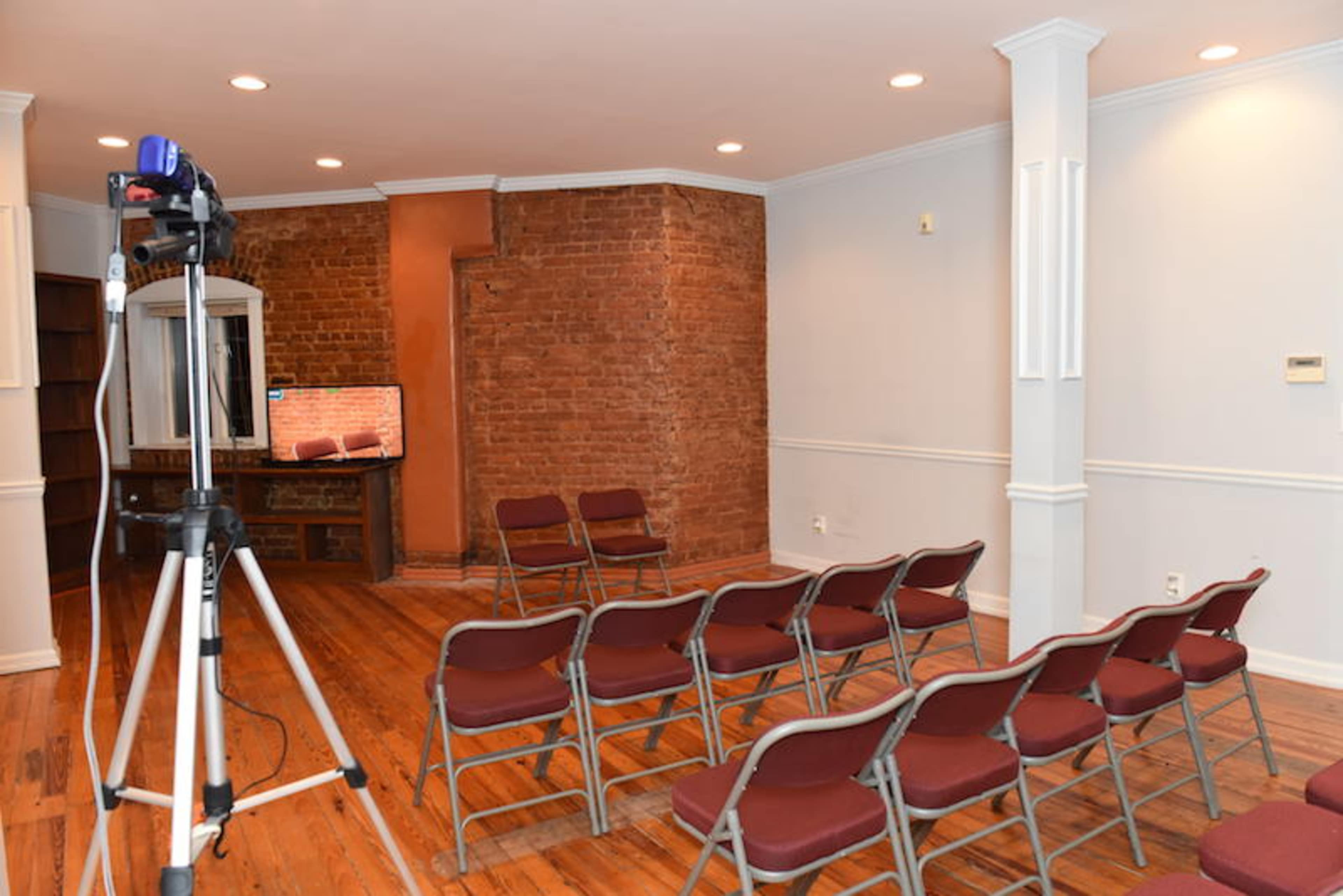 A small room with wooden floors, featuring several chairs arranged in rows facing a television and a brick wall.