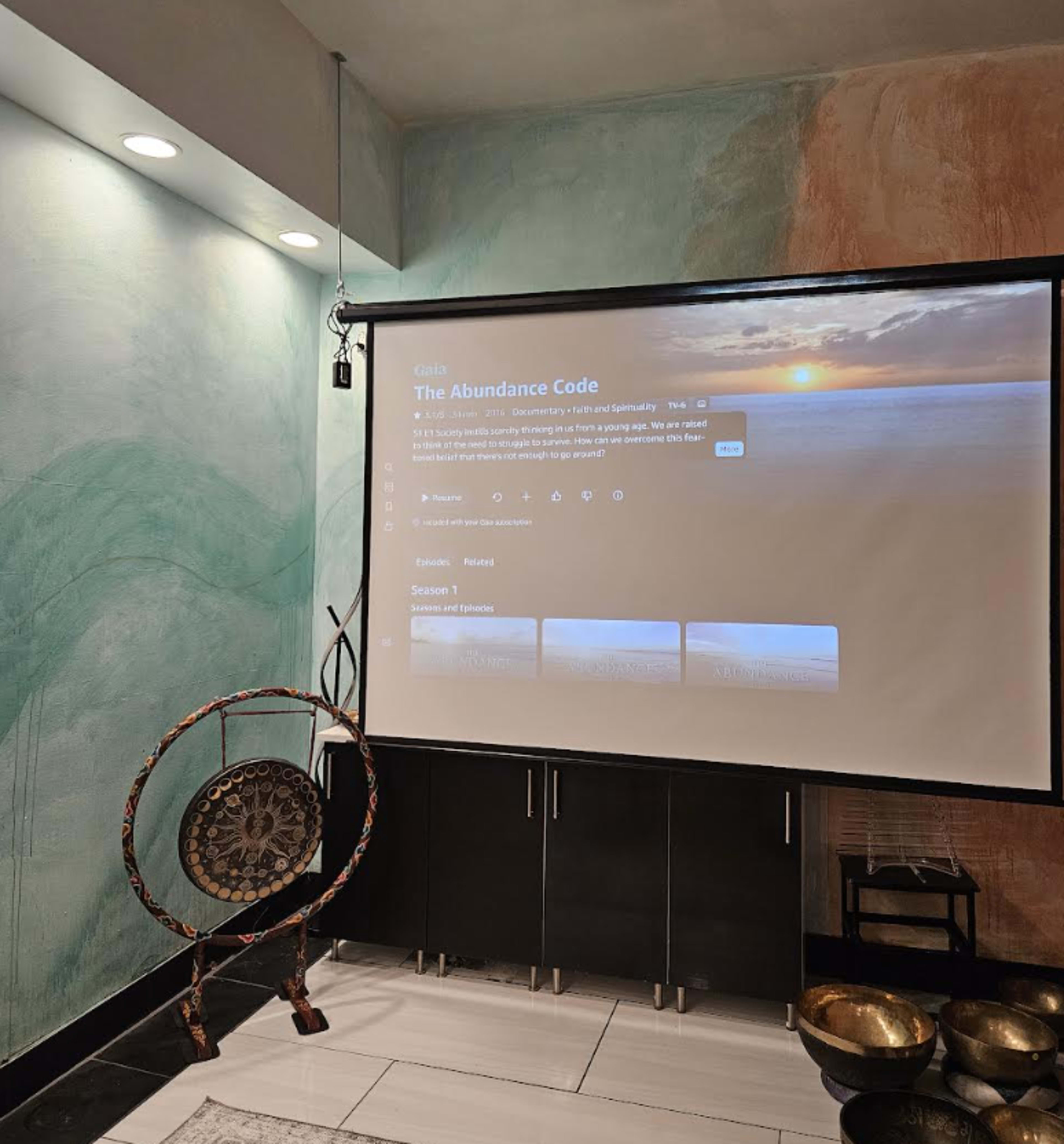 A large screen displays the title "The Abundance Code" against a softly colored wall, with a gong and various bowls positioned nearby.