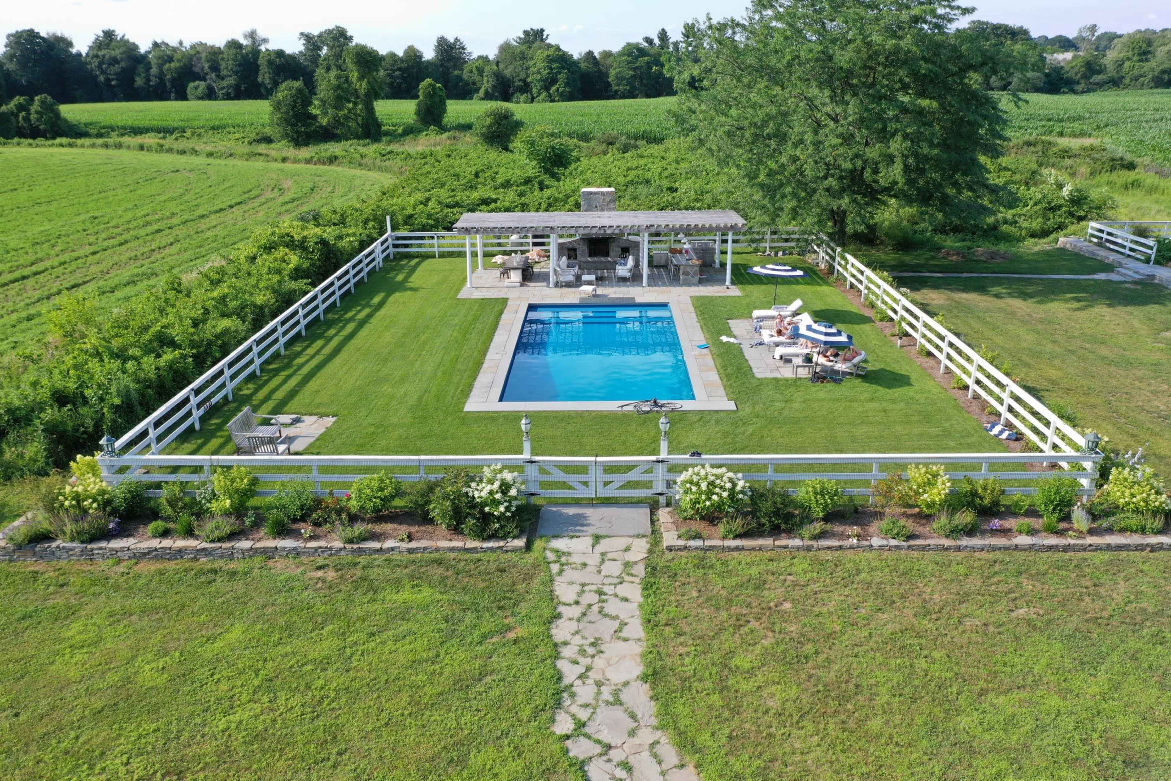 A fenced-in backyard with a swimming pool, a gazebo, and landscaped greenery surrounded by a large field.