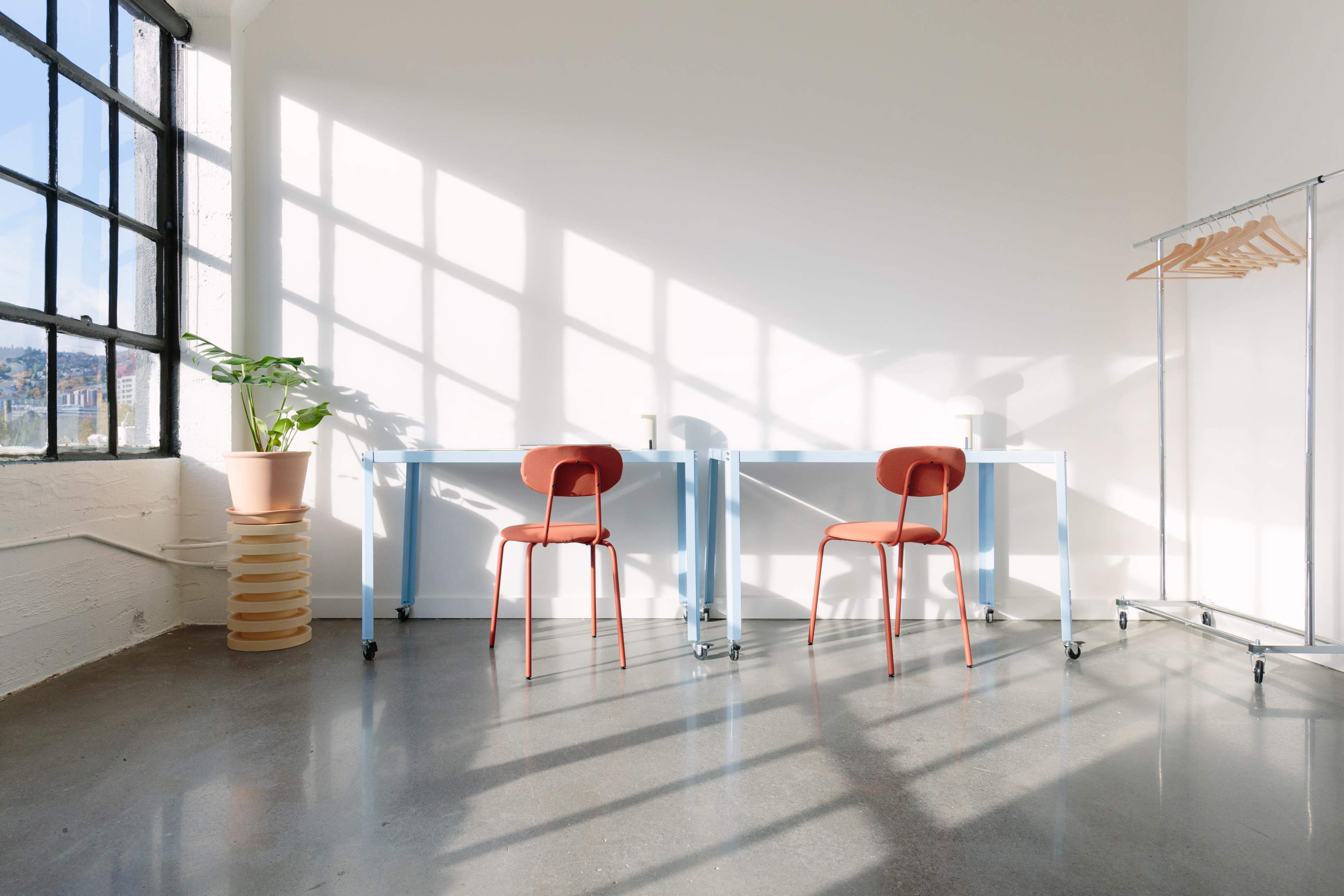 A bright, minimalist room with two red chairs at a blue table, large windows casting shadows on the polished concrete floor, and a plant next to a clothing rack.