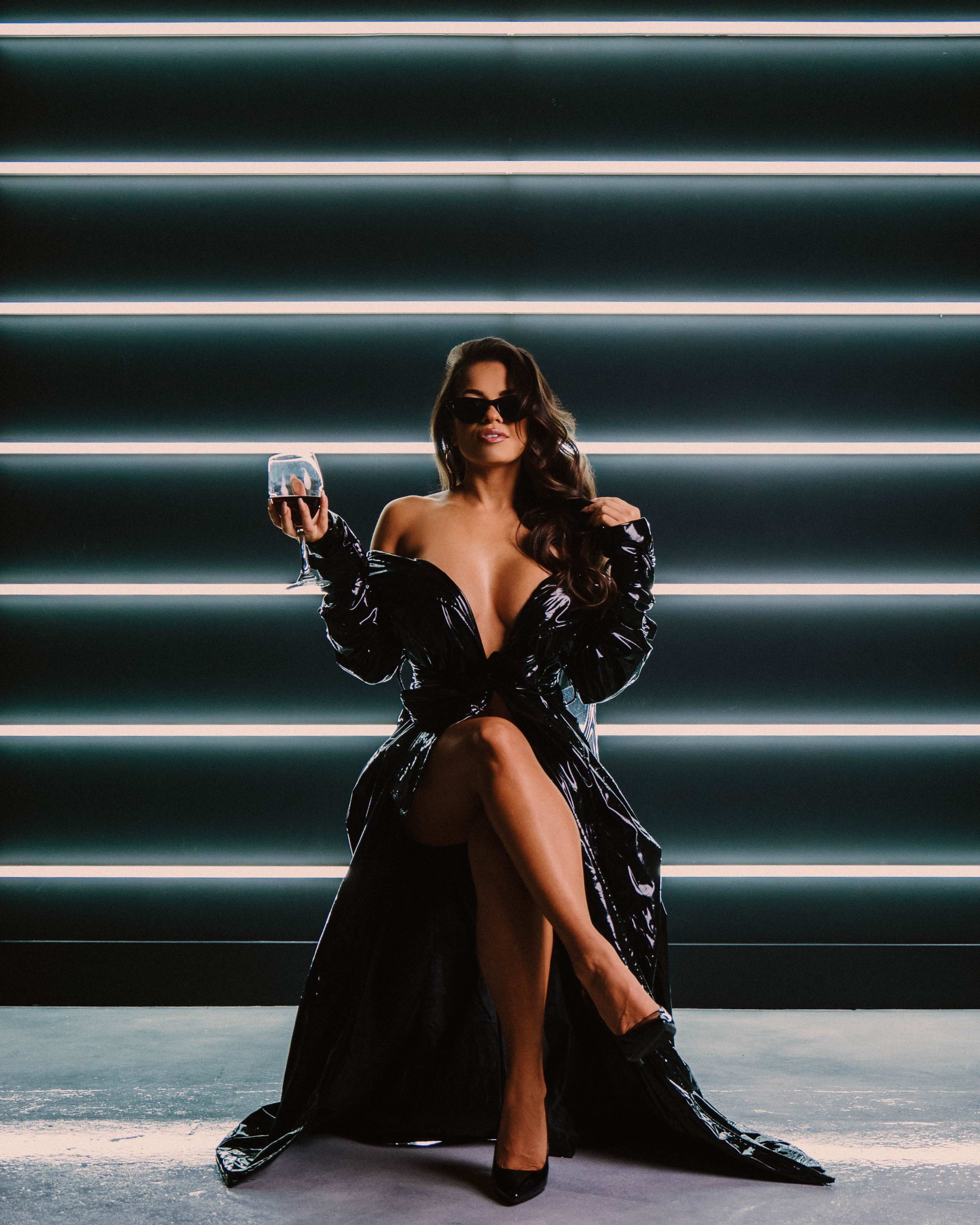 A woman in a black, glossy dress holds a wine glass while seated against a backdrop of vertical neon lights.