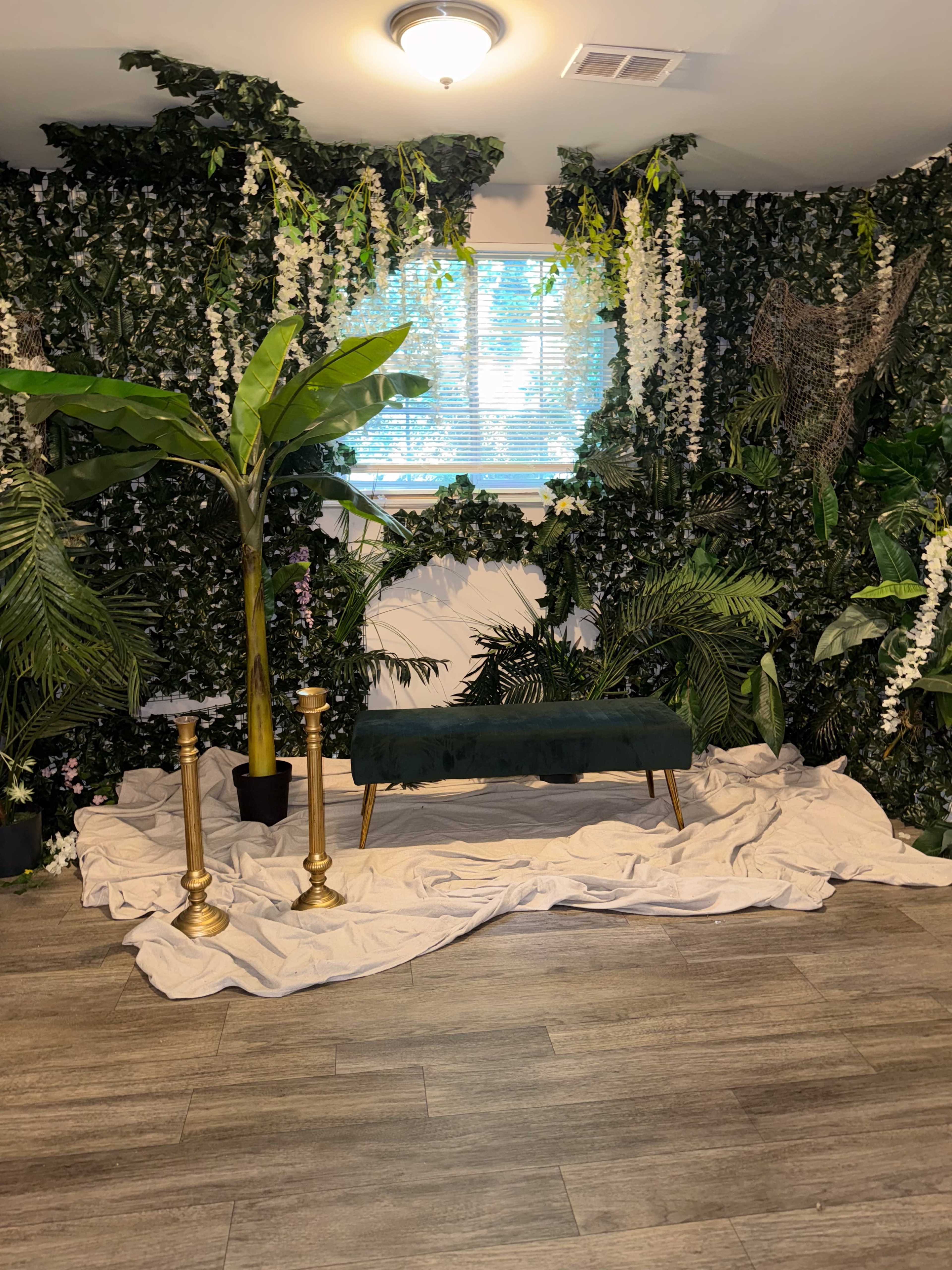 The scene features a green velvet bench surrounded by a wall of lush greenery, with decorative plants and a light-colored cloth on the floor.