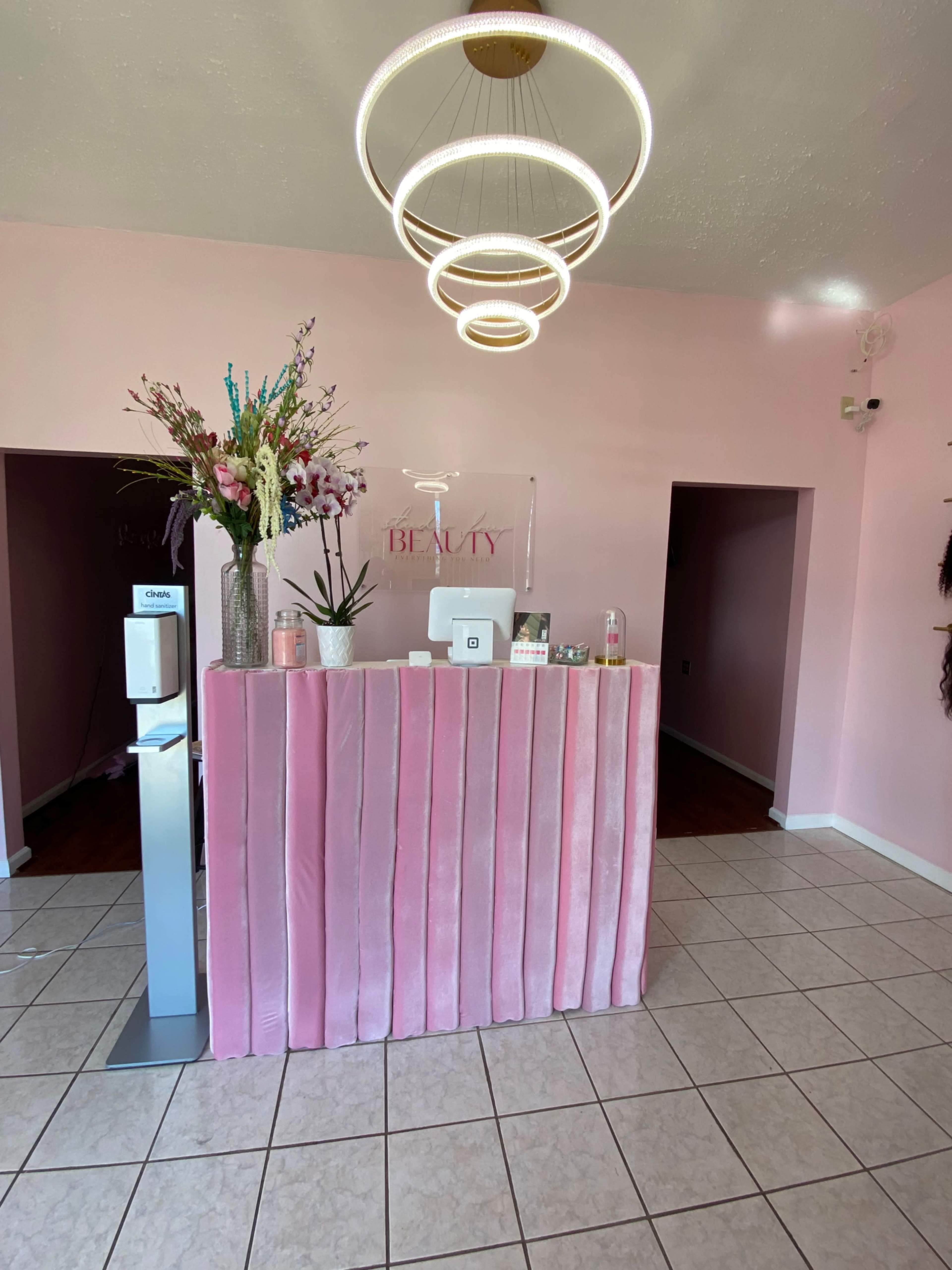 A beauty salon reception area with a pink velvet front desk, decorative flowers, and modern circular light fixtures hanging from the ceiling.