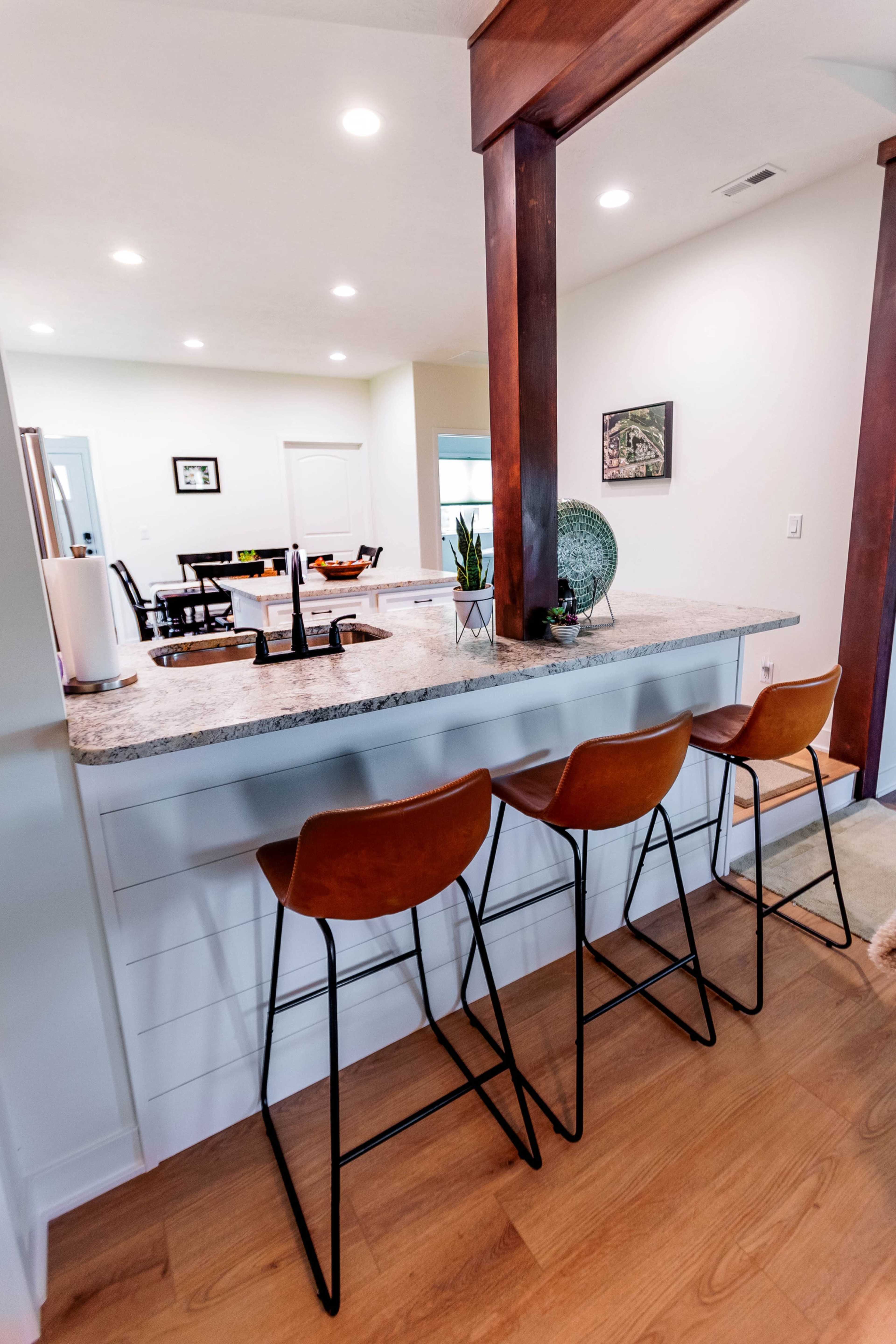 A modern kitchen features a bar counter with three leather stools, illuminated by recessed lighting.