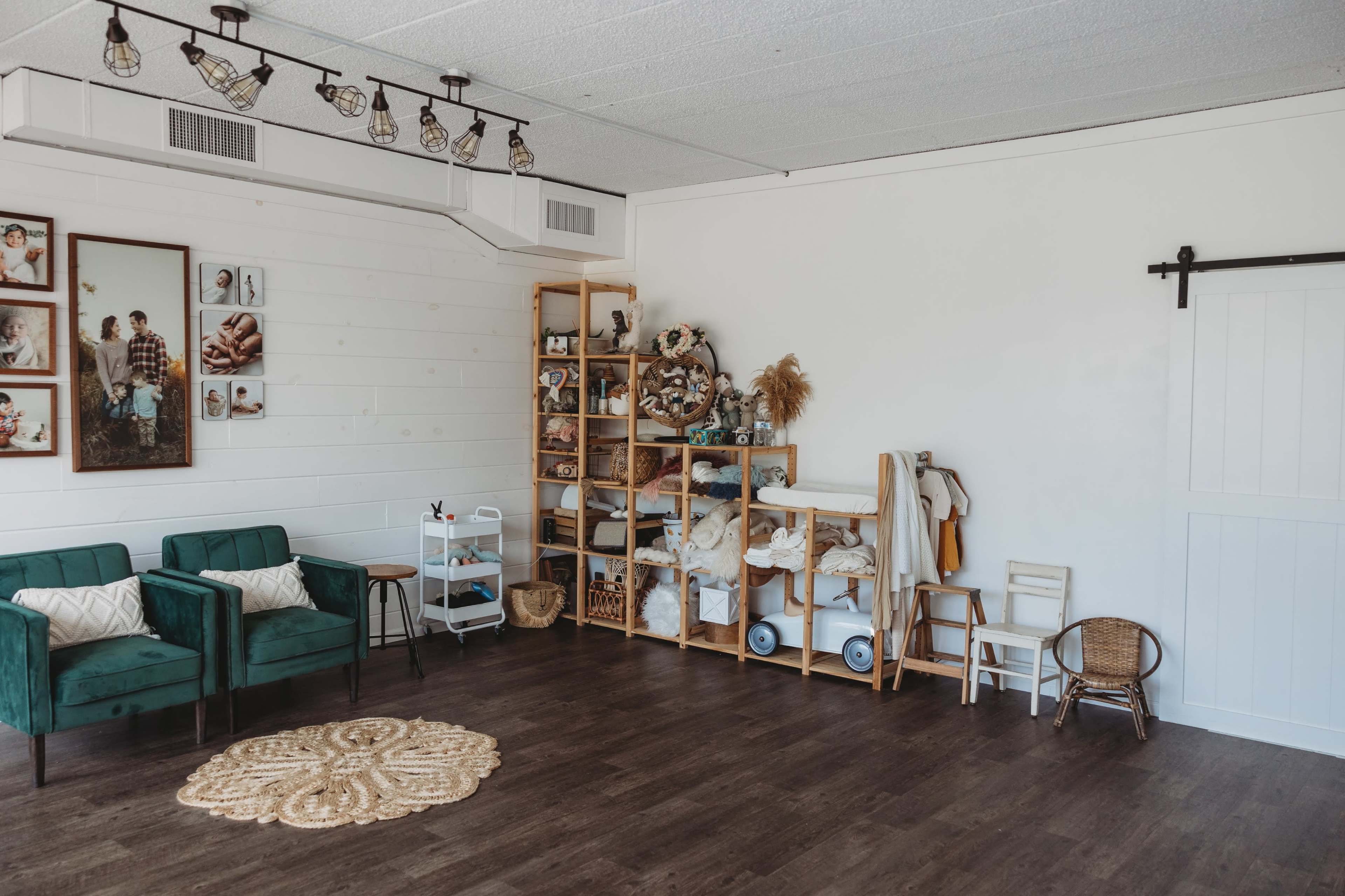 A spacious room with two green chairs, wooden shelving filled with various items, and a small round rug on the dark wood floor.
