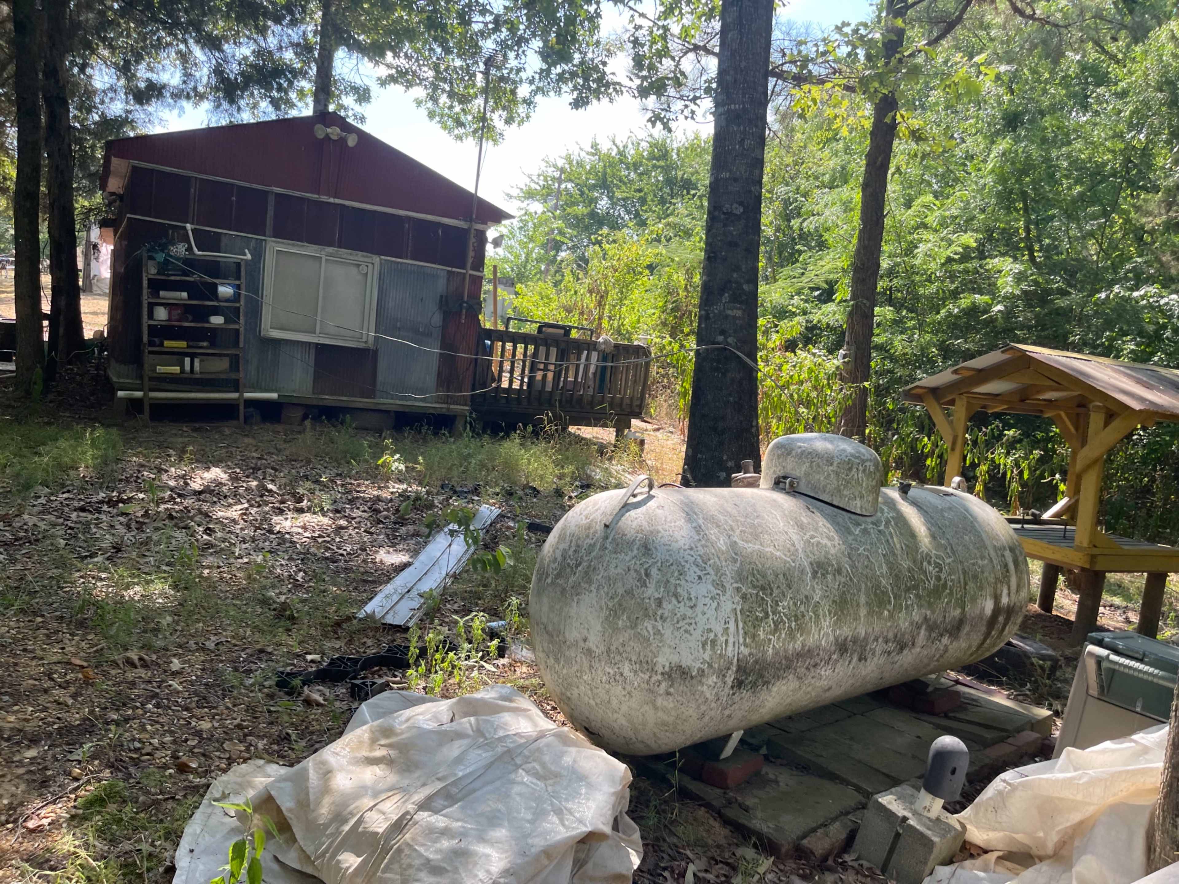 A propane tank sits on a concrete base in front of a red barn-like structure surrounded by trees.