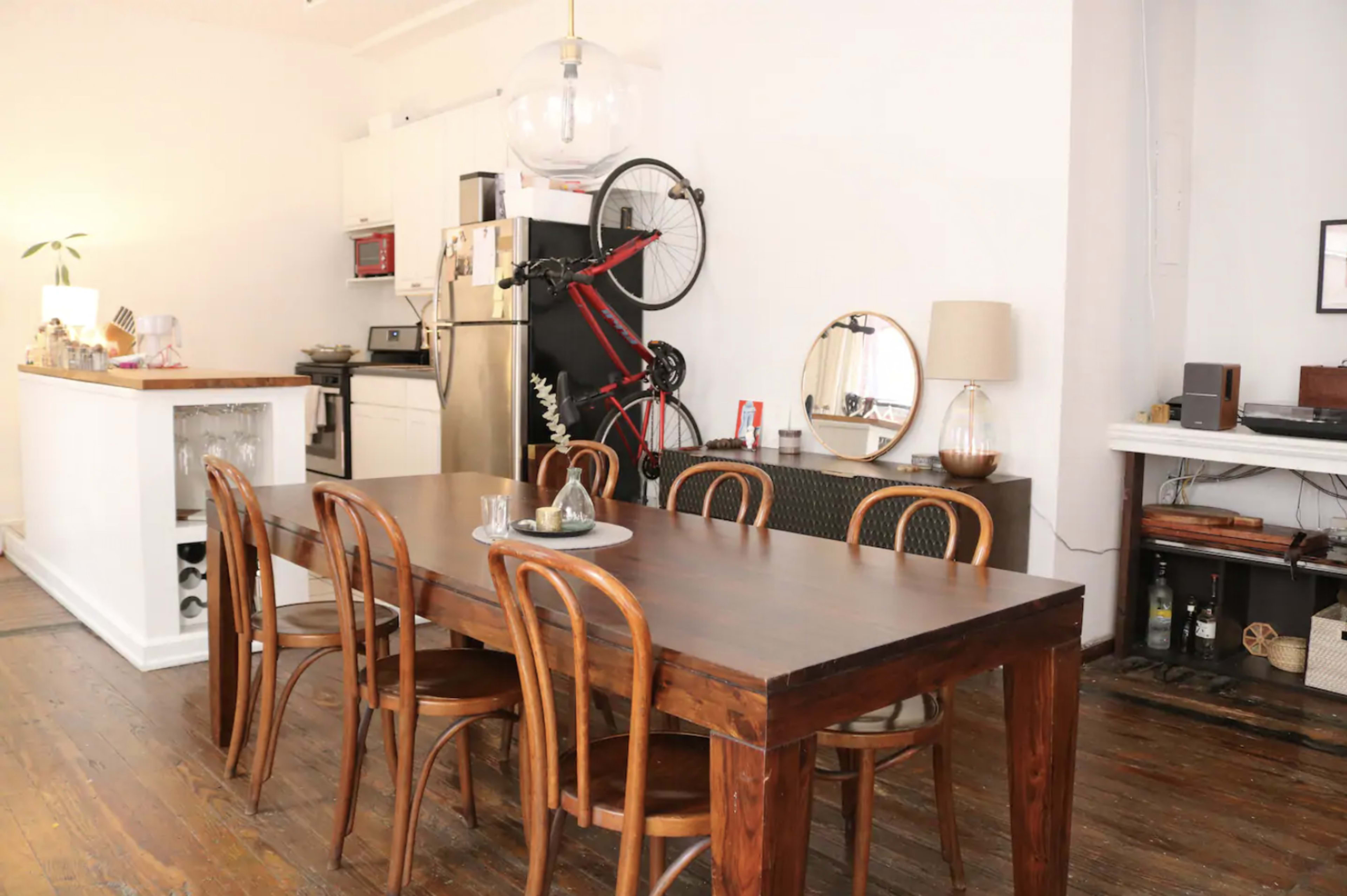 A spacious, well-lit dining area features a long wooden table with curved-back chairs, against a backdrop of a minimalist kitchen and a bicycle mounted on the wall.
