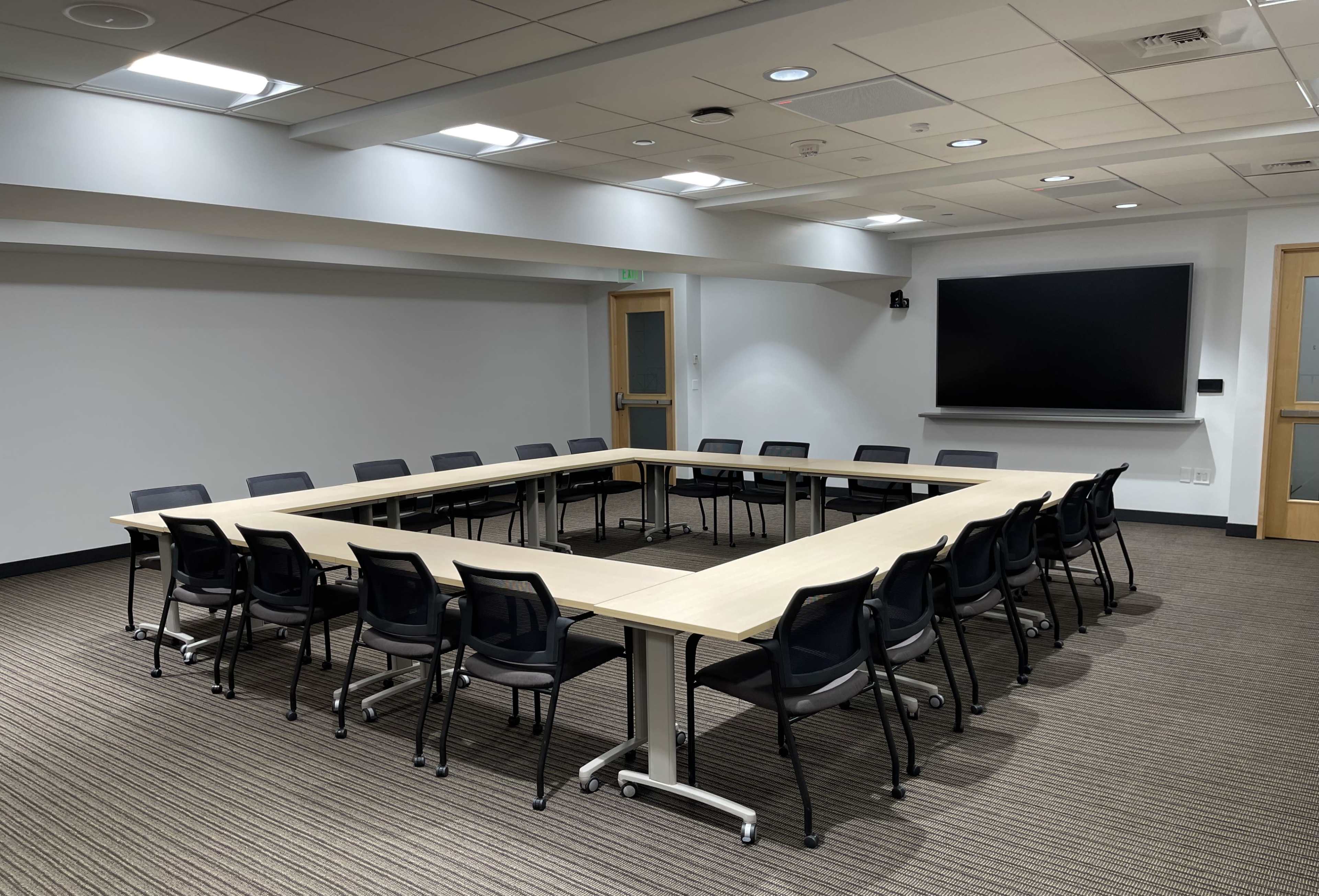 A conference room features a large rectangular table surrounded by black chairs, with white walls, a dark screen, and overhead lights.