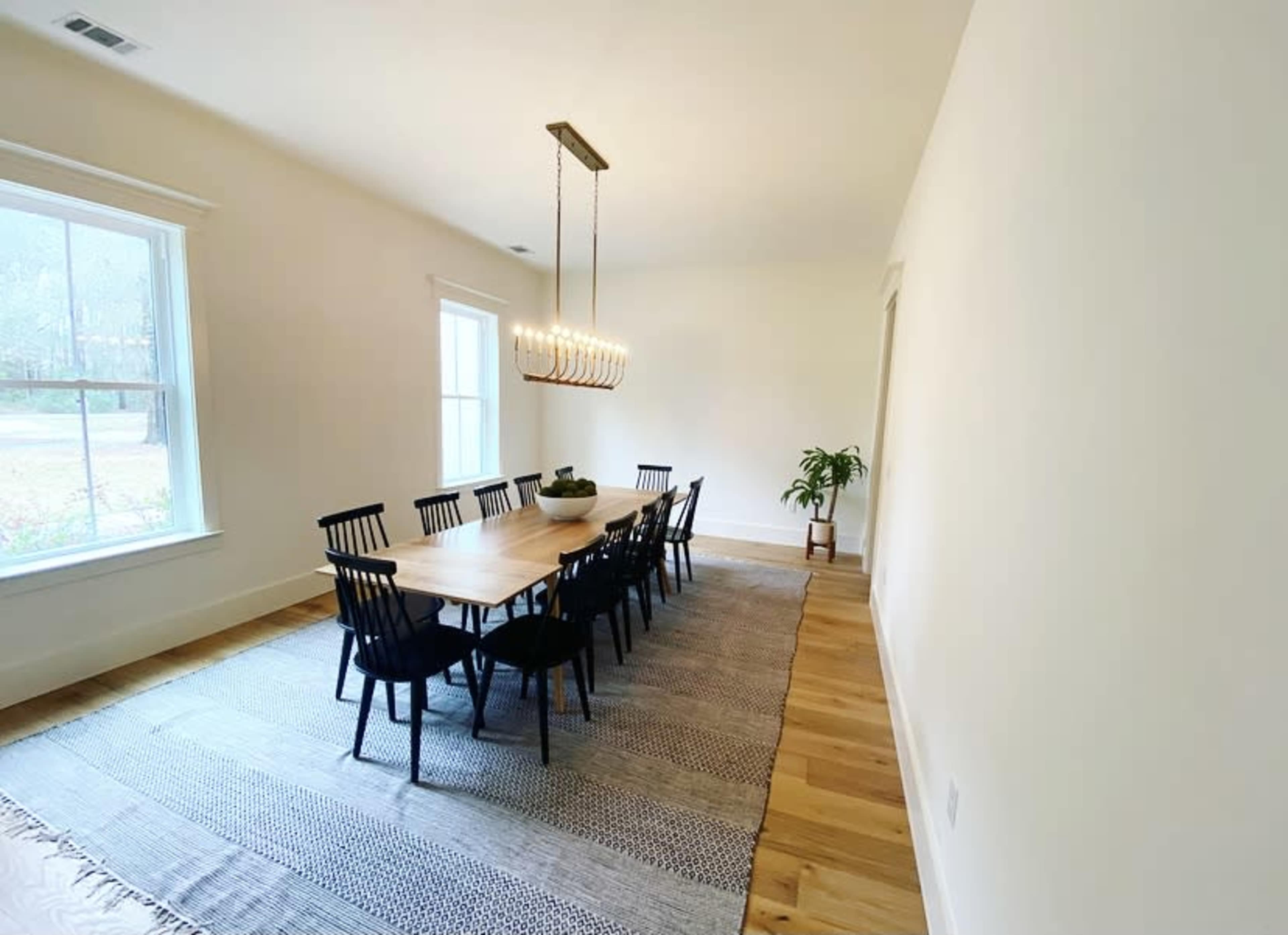 A spacious dining room features a long wooden table surrounded by black chairs, with a chandelier overhead and a potted plant in the corner.