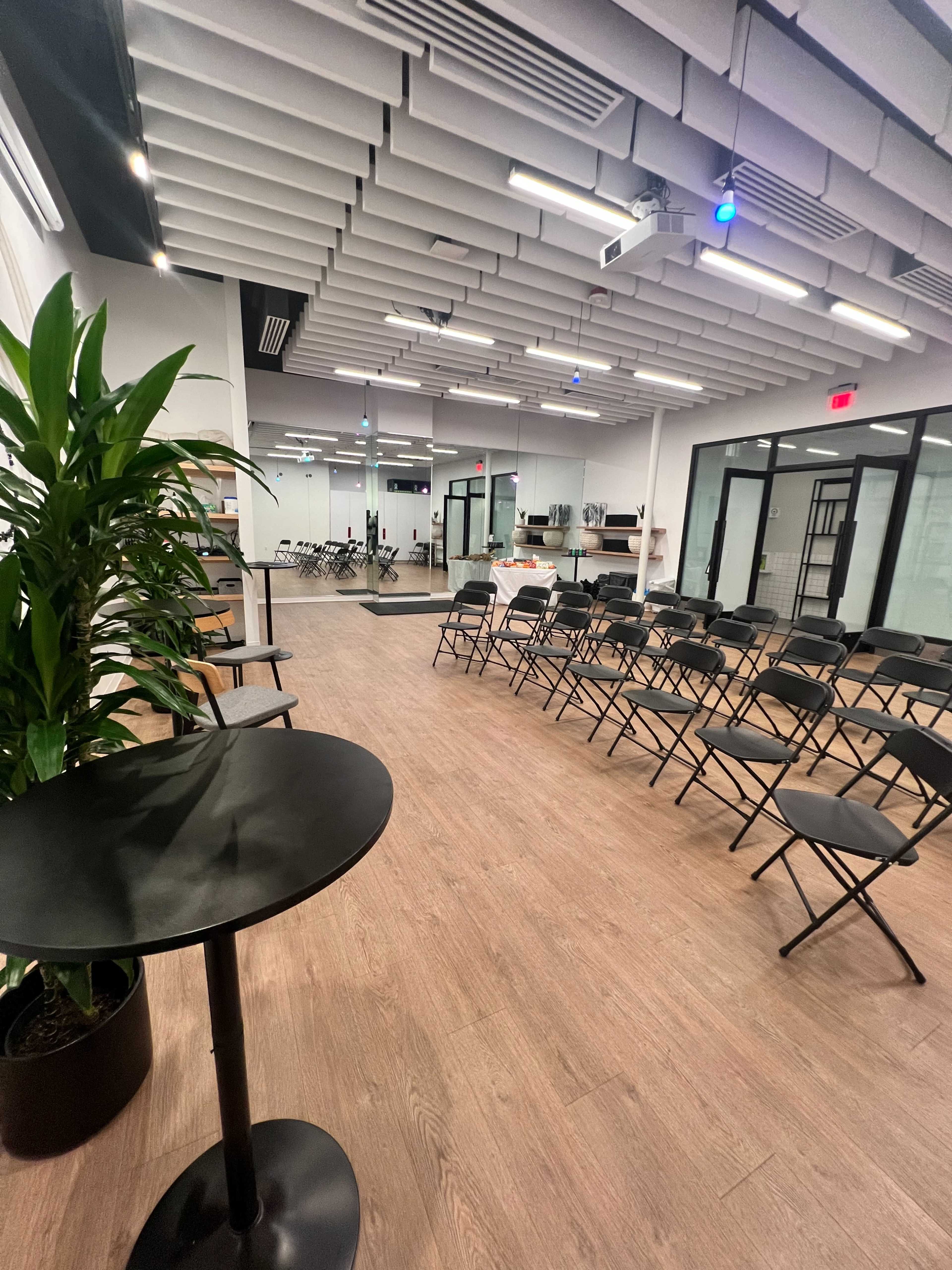 A spacious, modern meeting room with black folding chairs arranged in rows and a small round table with a plant beside it.