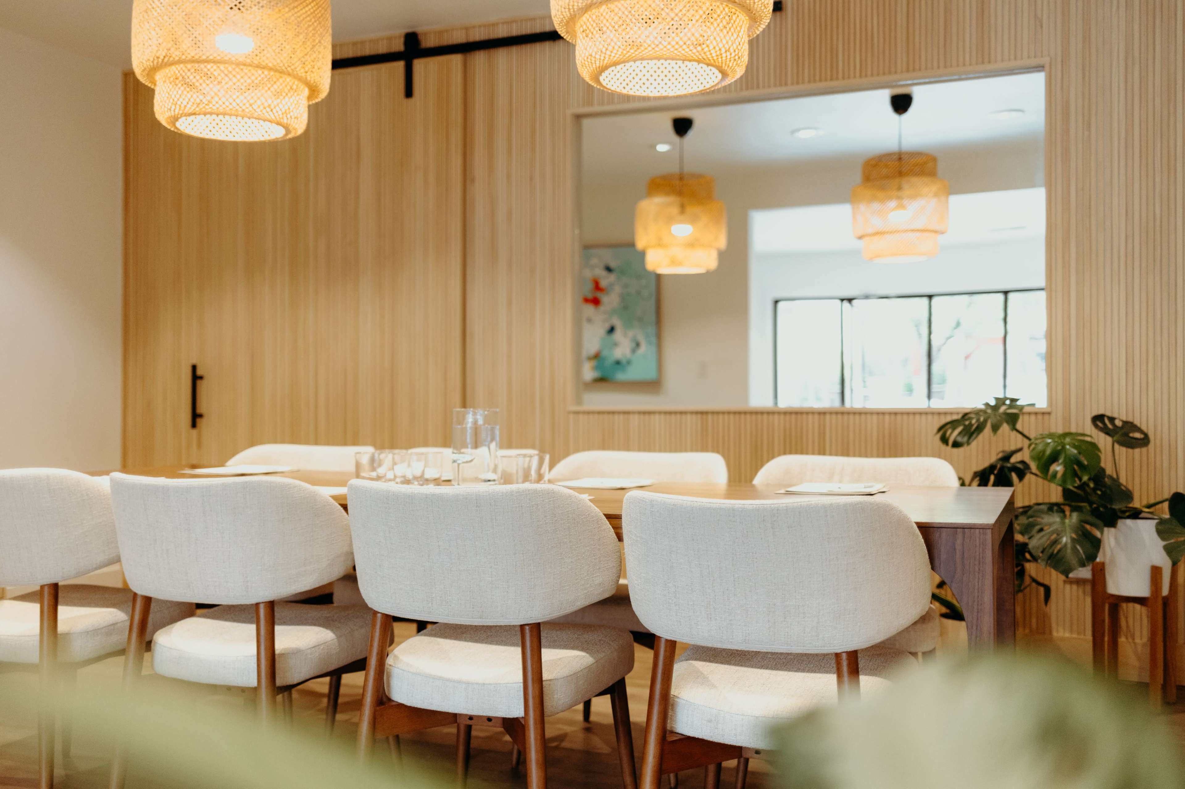 A wooden dining table is set with glassware, surrounded by upholstered chairs, beneath pendant lights, with a large mirror reflecting the space.