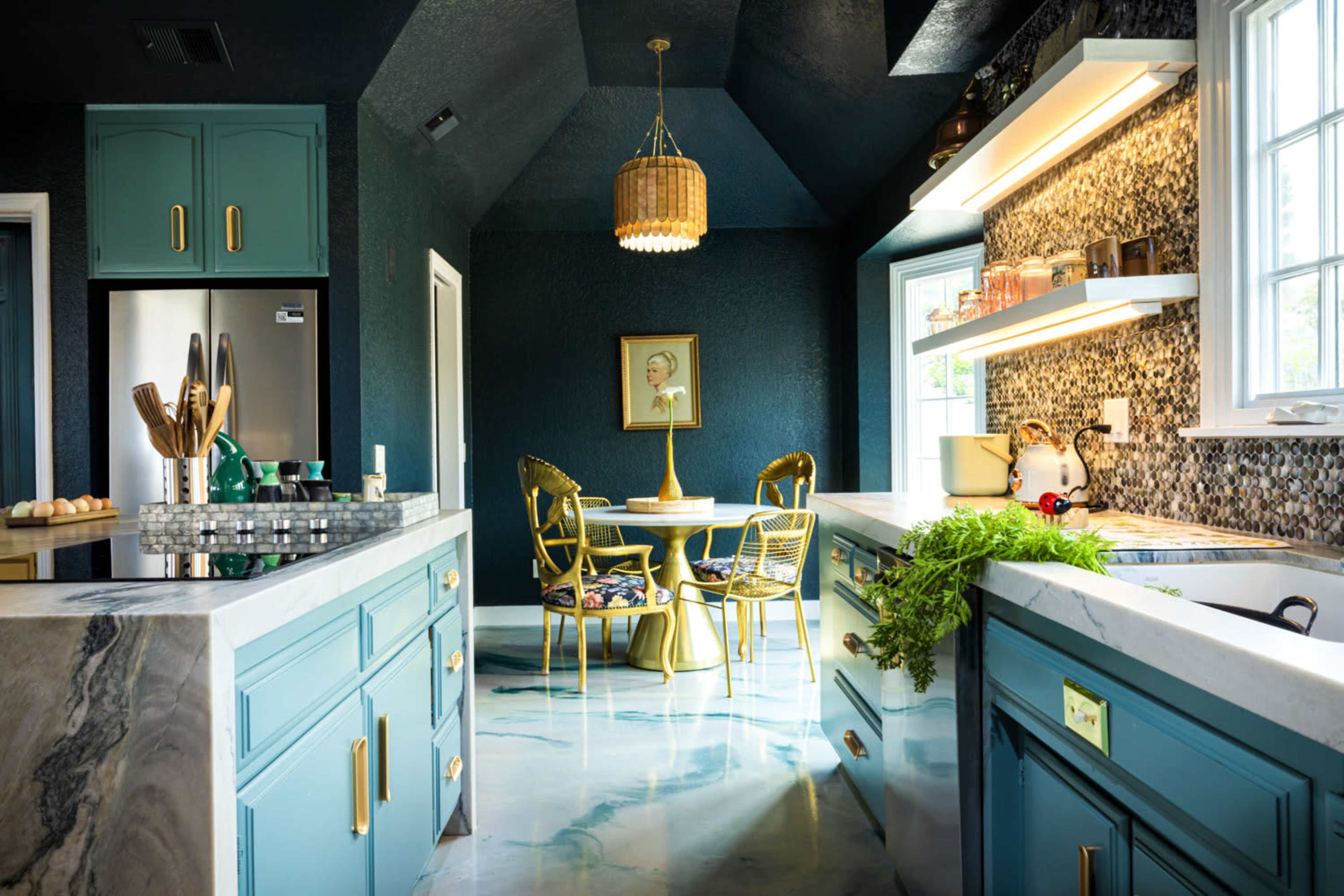 The image shows a modern kitchen with teal cabinets, a marble countertop, and a small dining area featuring a round table and four chairs, all set against a dark blue wall and illuminated by overhead lighting.