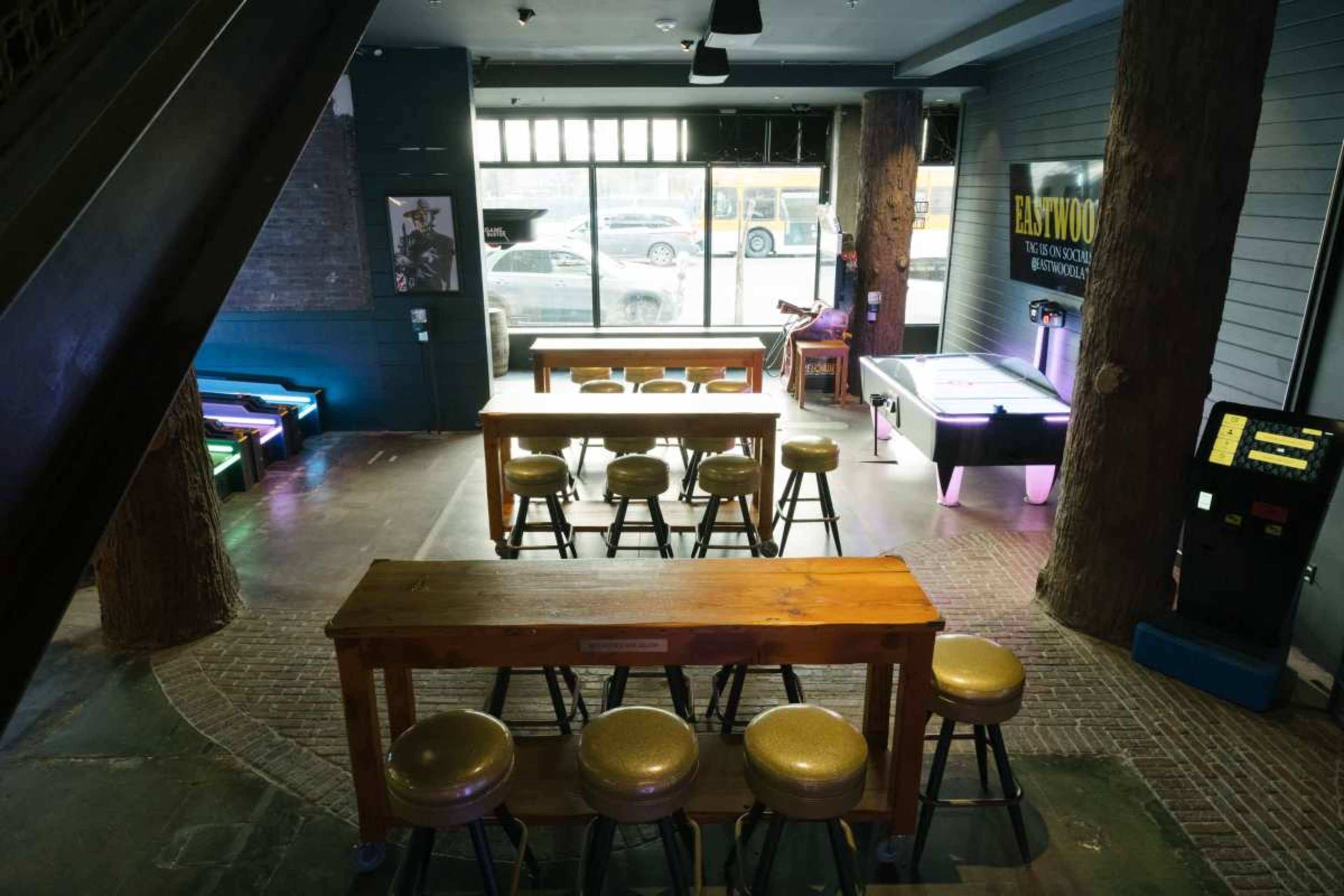 The image shows a bar area with wooden tables and stools, an air hockey table, and a vintage arcade game, all set in a dimly lit venue with large windows.
