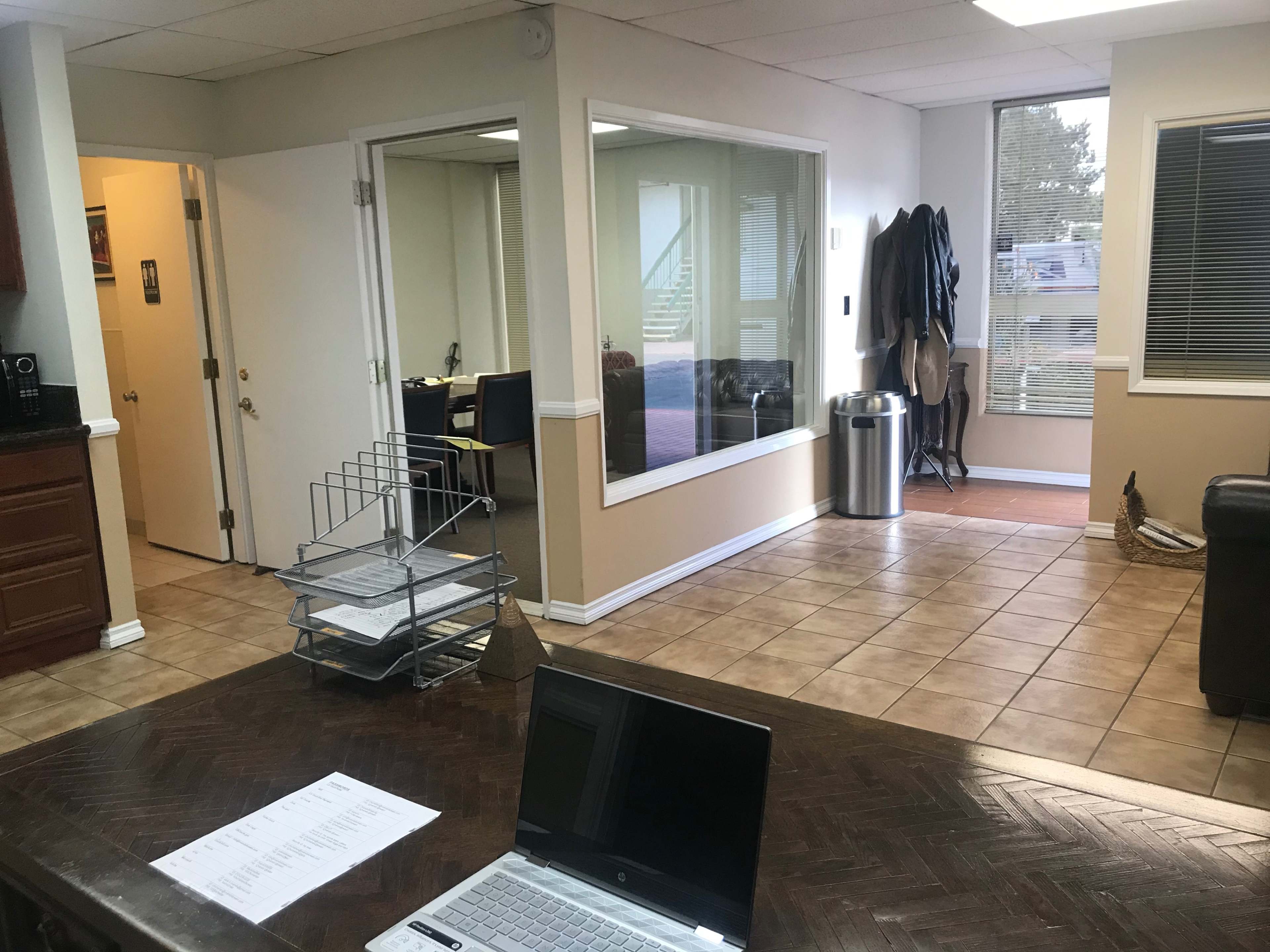 An office space with a reception area, a desk with a laptop, and a large window overlooking another room with chairs.