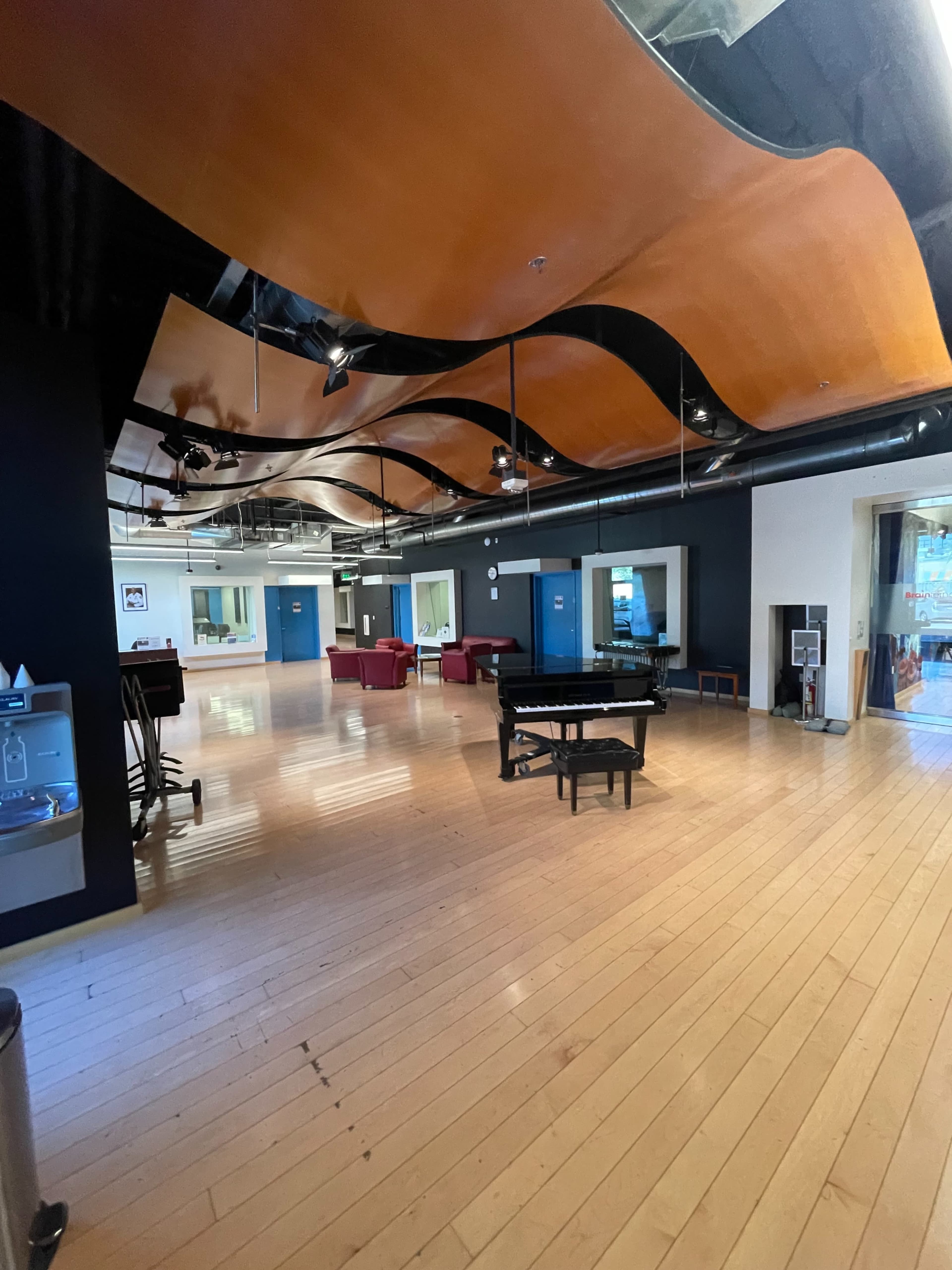 The image shows a spacious, well-lit interior room featuring wooden floors, a grand piano in the center, and a unique wavy ceiling design.