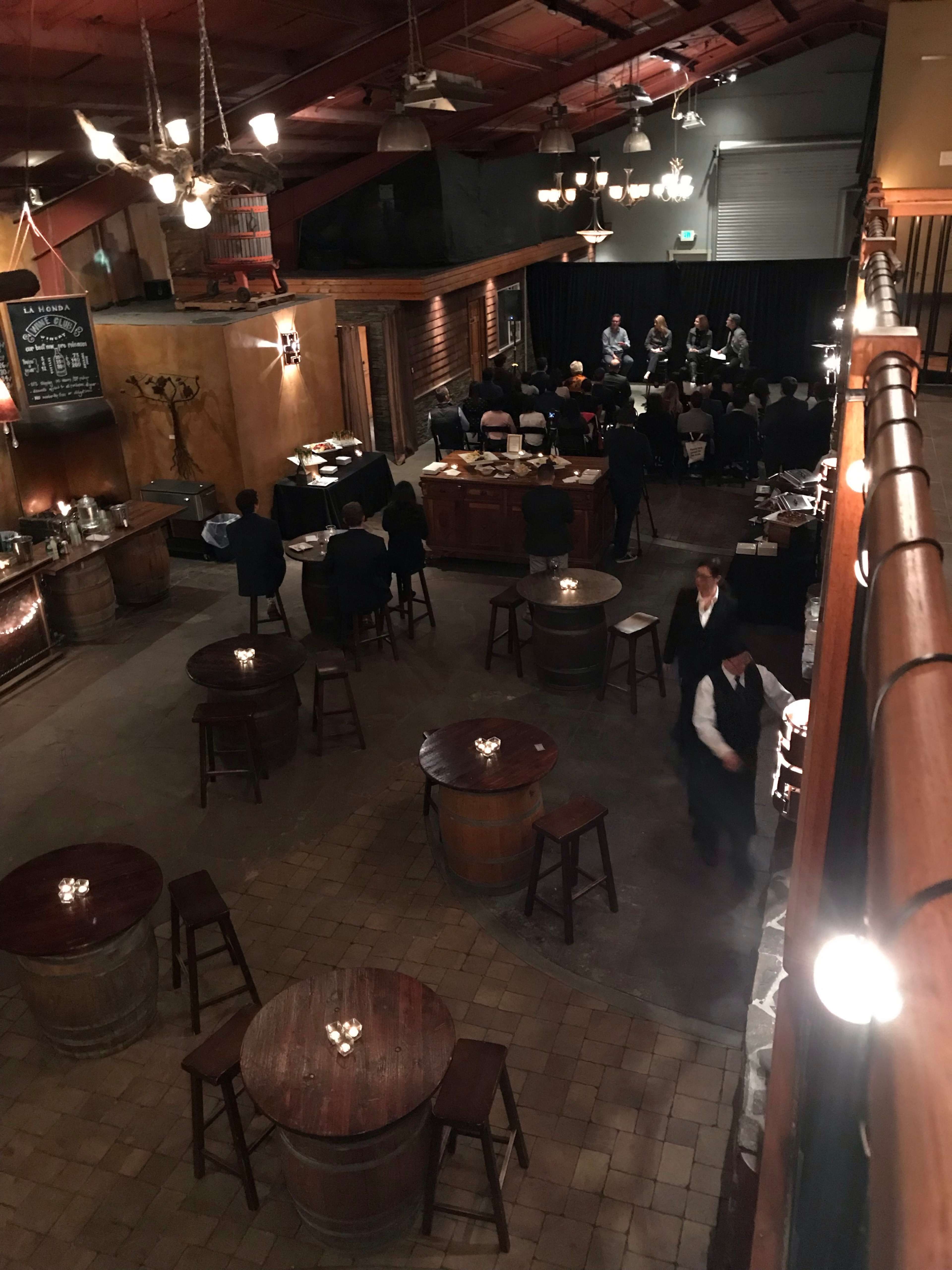 The image shows a dimly lit event space with round tables and bar stools, where a group is seated on a stage at the far end.