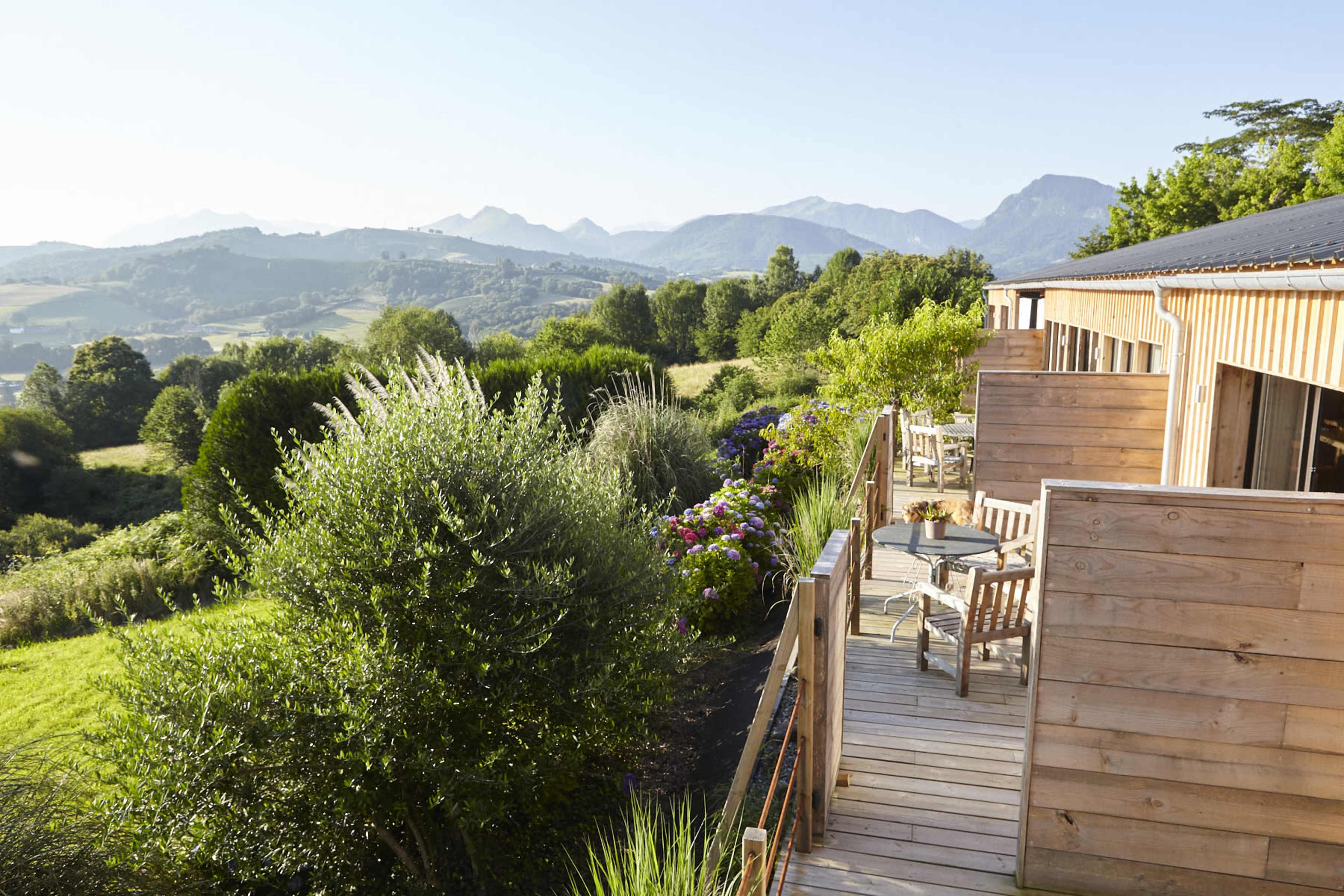 A wooden deck overlooks a lush landscape with mountains in the background and a variety of plants and flowers in the foreground.