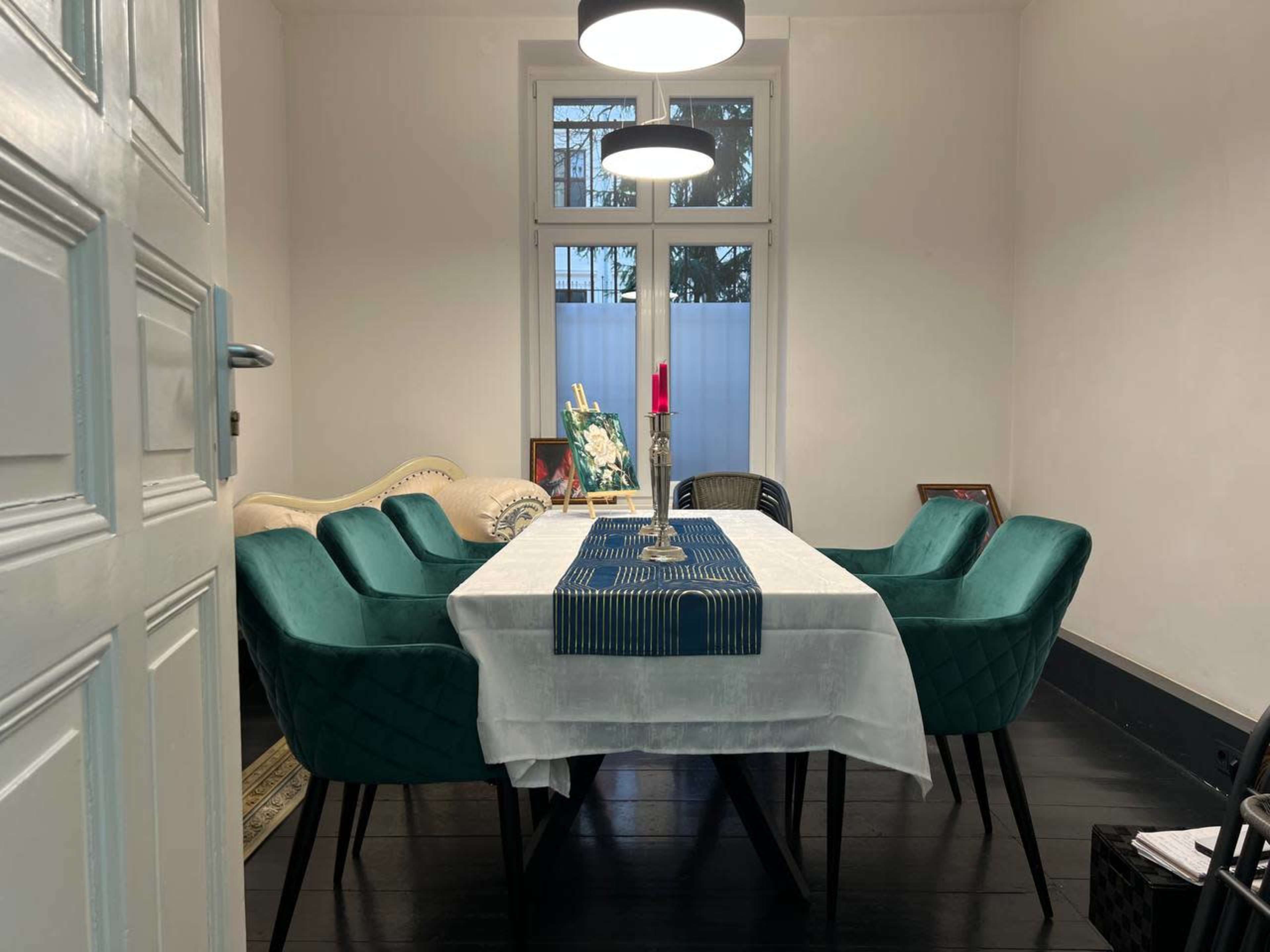 A dining room features a rectangular table set with a white tablecloth and a blue runner, surrounded by six teal chairs, and a candle holder with a red candle placed in the center.