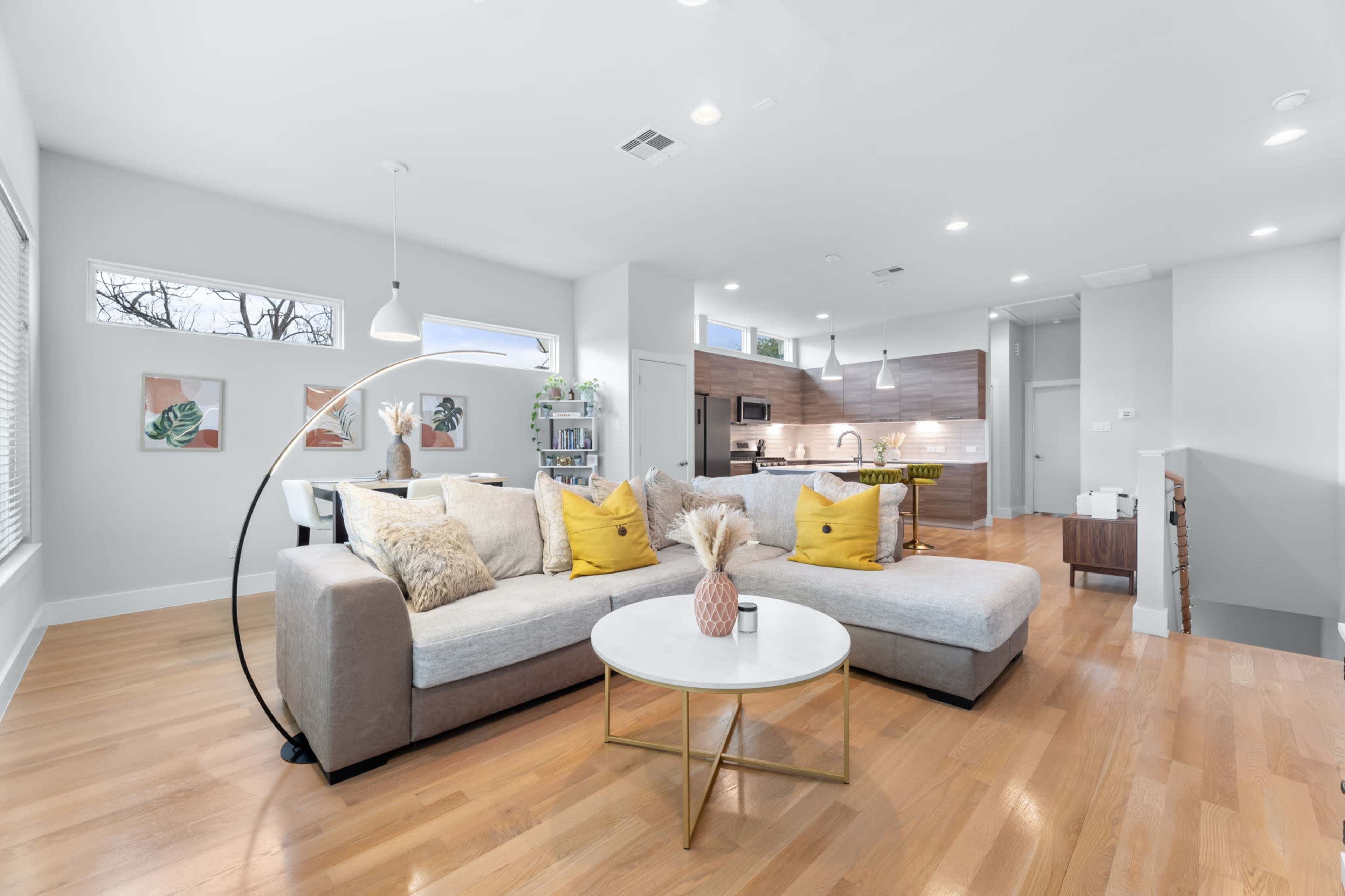 A modern living room features a gray sectional sofa with yellow accent pillows and a round coffee table, leading into a kitchen with wooden cabinetry.