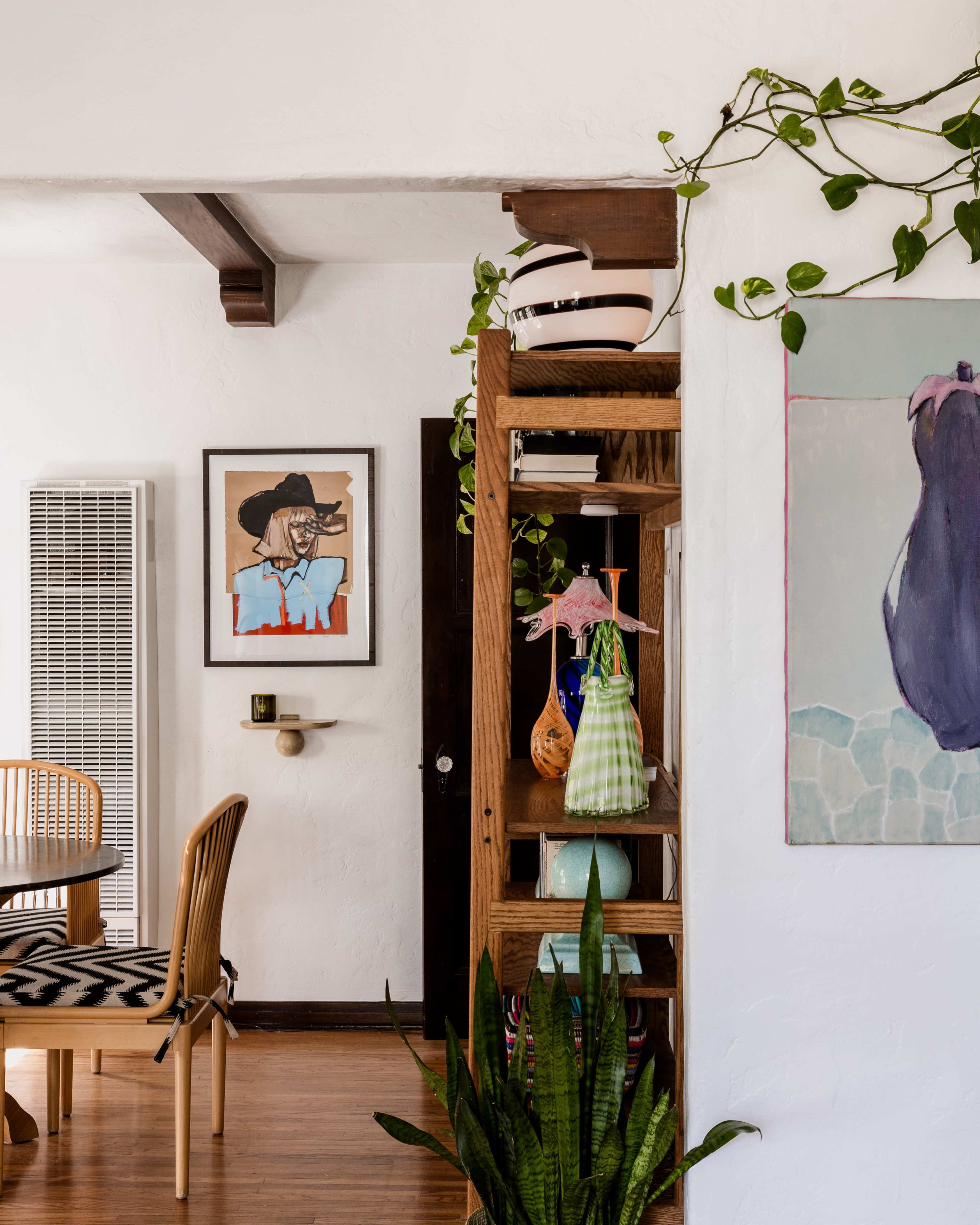 The image shows a cozy interior space featuring a wooden bookshelf with decorative items, an art piece on the wall, and a dining area in the background.