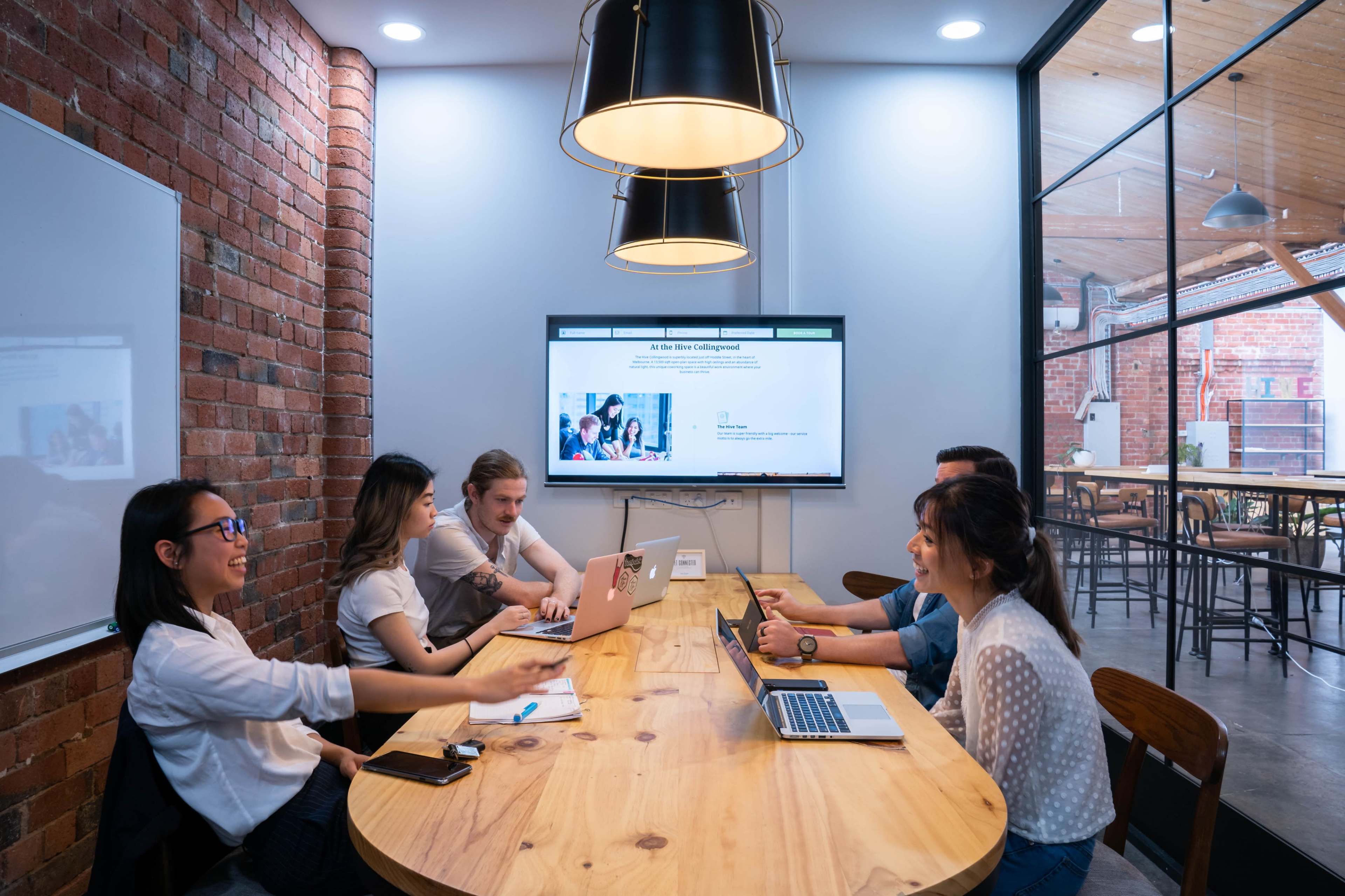 A group of five people are engaged in a meeting around a wooden table in a well-lit conference room with brick walls and a large screen displaying a presentation.