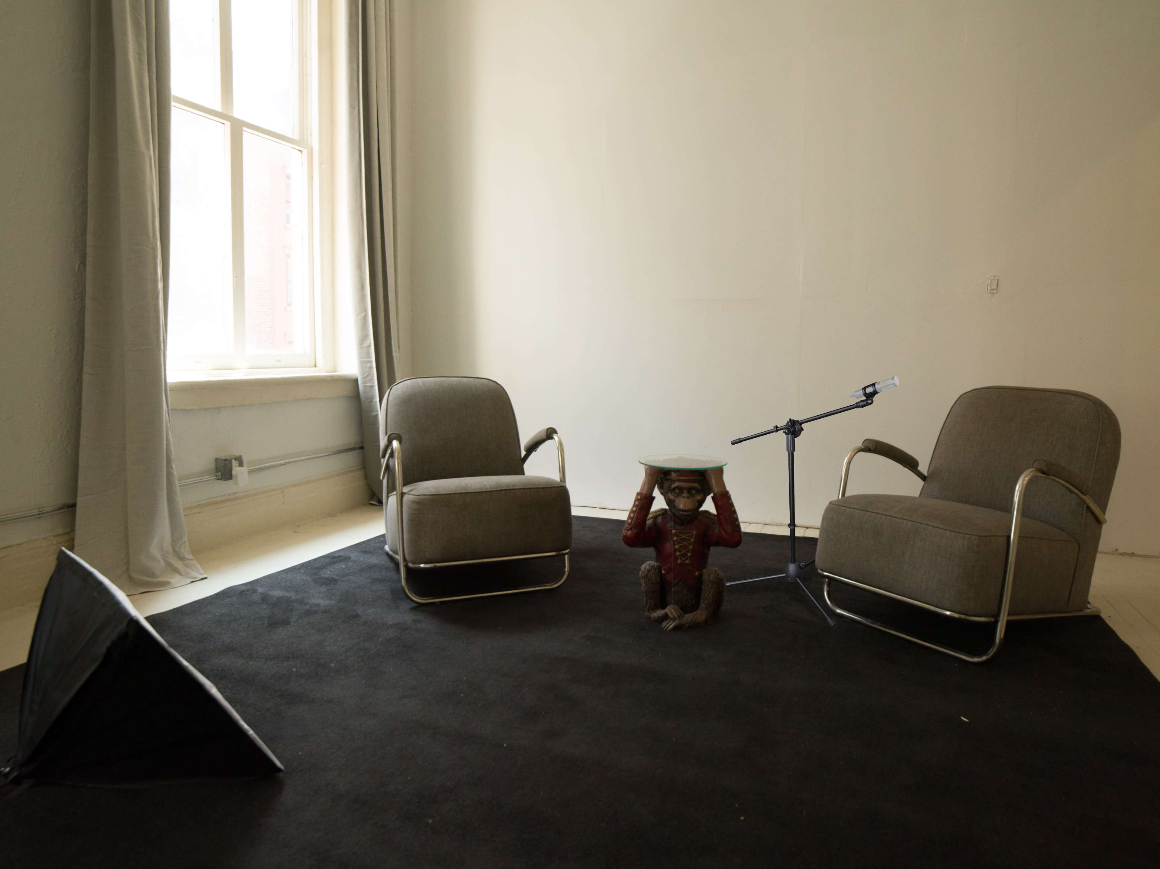The image shows two gray armchairs facing each other on a black carpet, with a small statue of a monkey holding a plate between them and a microphone on a stand nearby.