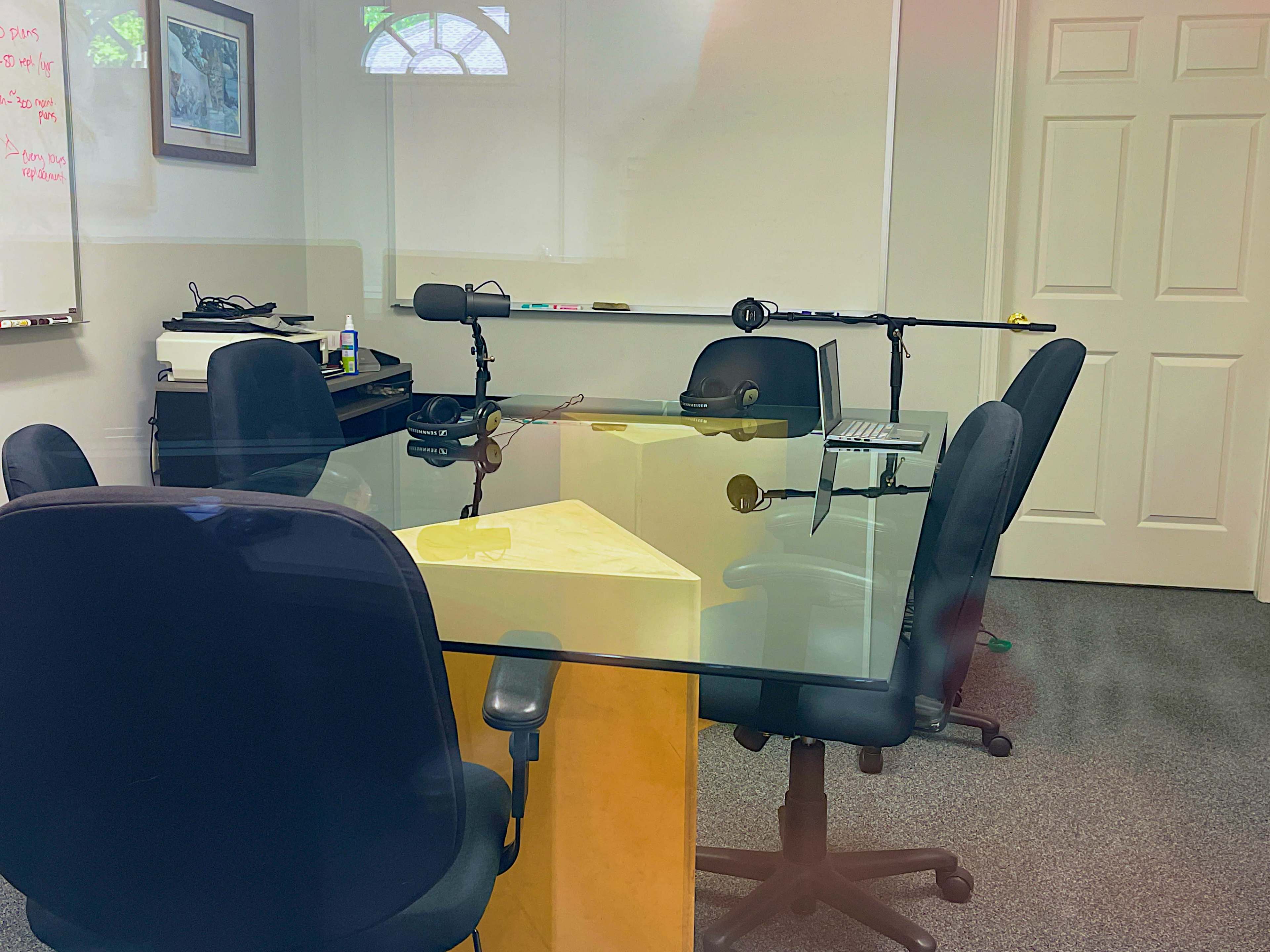 The image shows a conference room with a glass table, several black chairs, and equipment set up for audio recording.