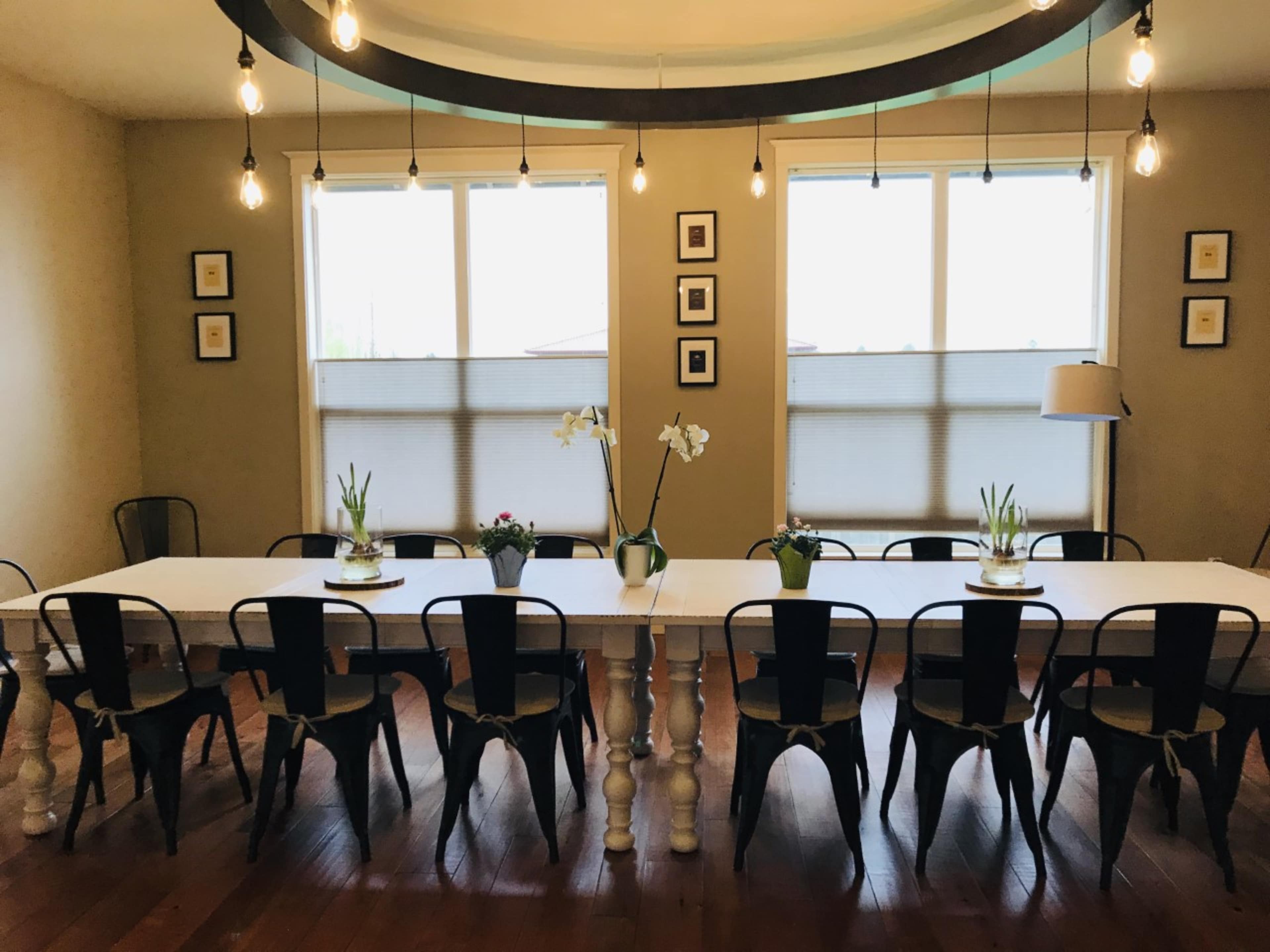 A long, white dining table with black chairs is set in a well-lit room featuring large windows and decorative plants.