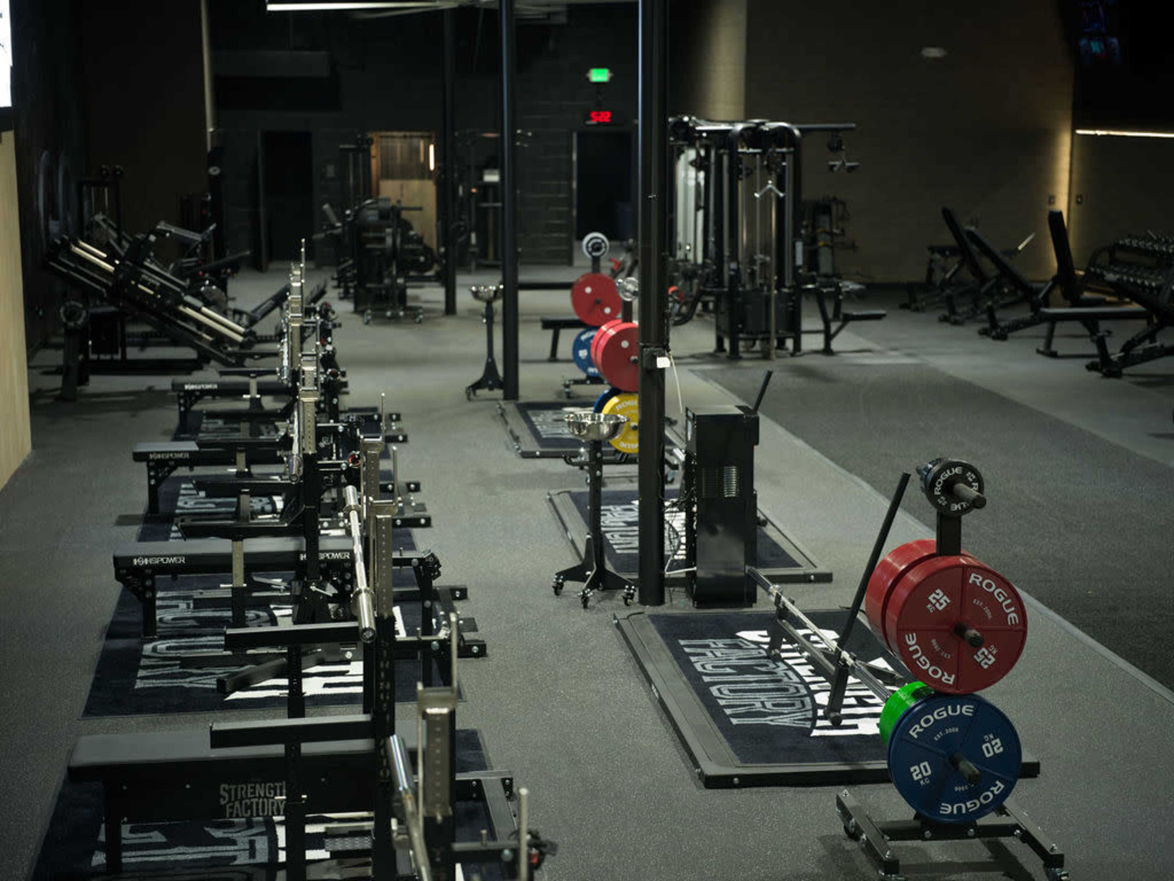 The image shows an empty gym interior equipped with various weightlifting machines, free weights, and fitness equipment arranged neatly on a rubberized floor.