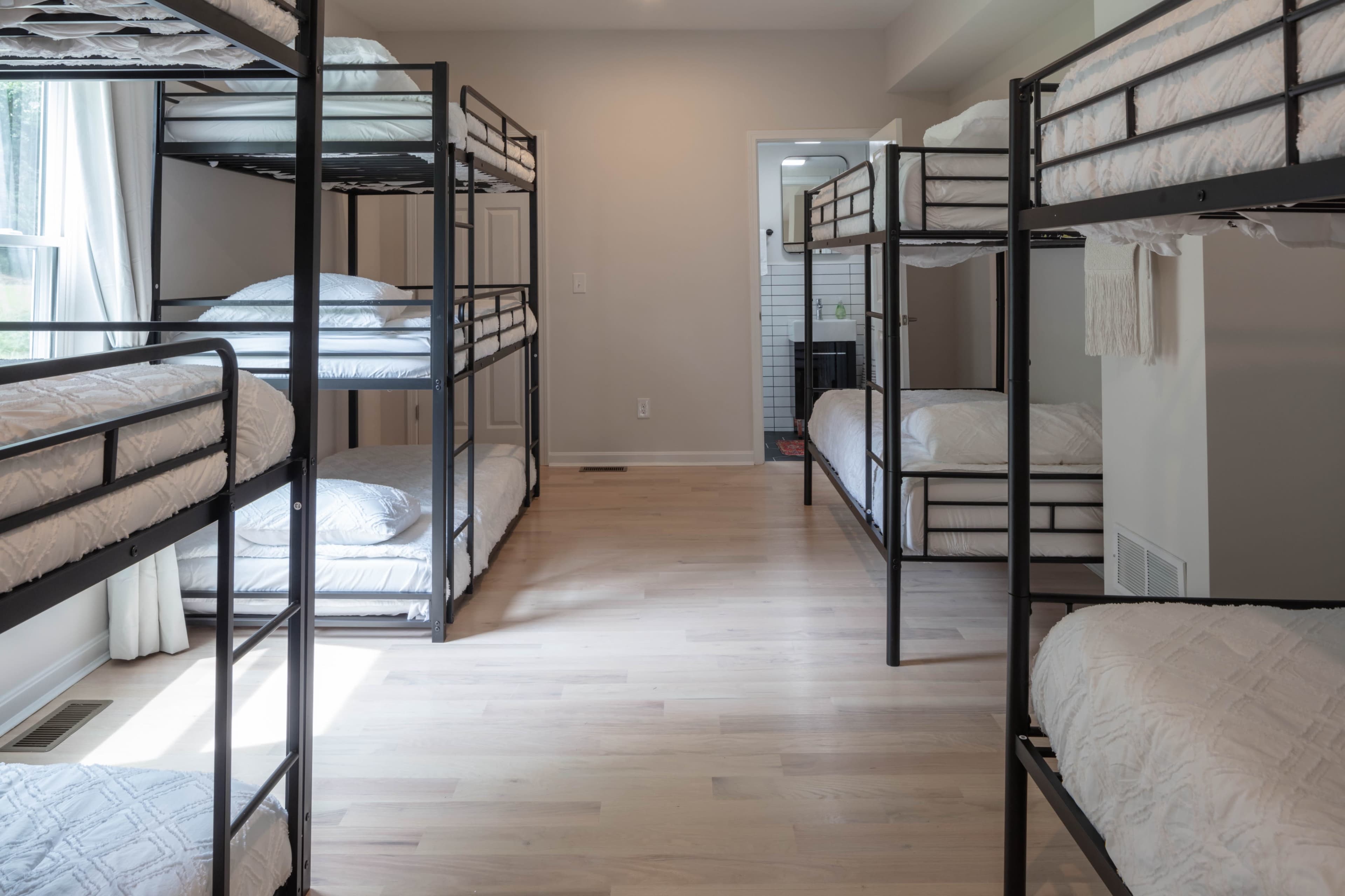 The image shows a room with several metal bunk beds arranged around a wooden floor, with white bedding on the top and bottom bunks.
