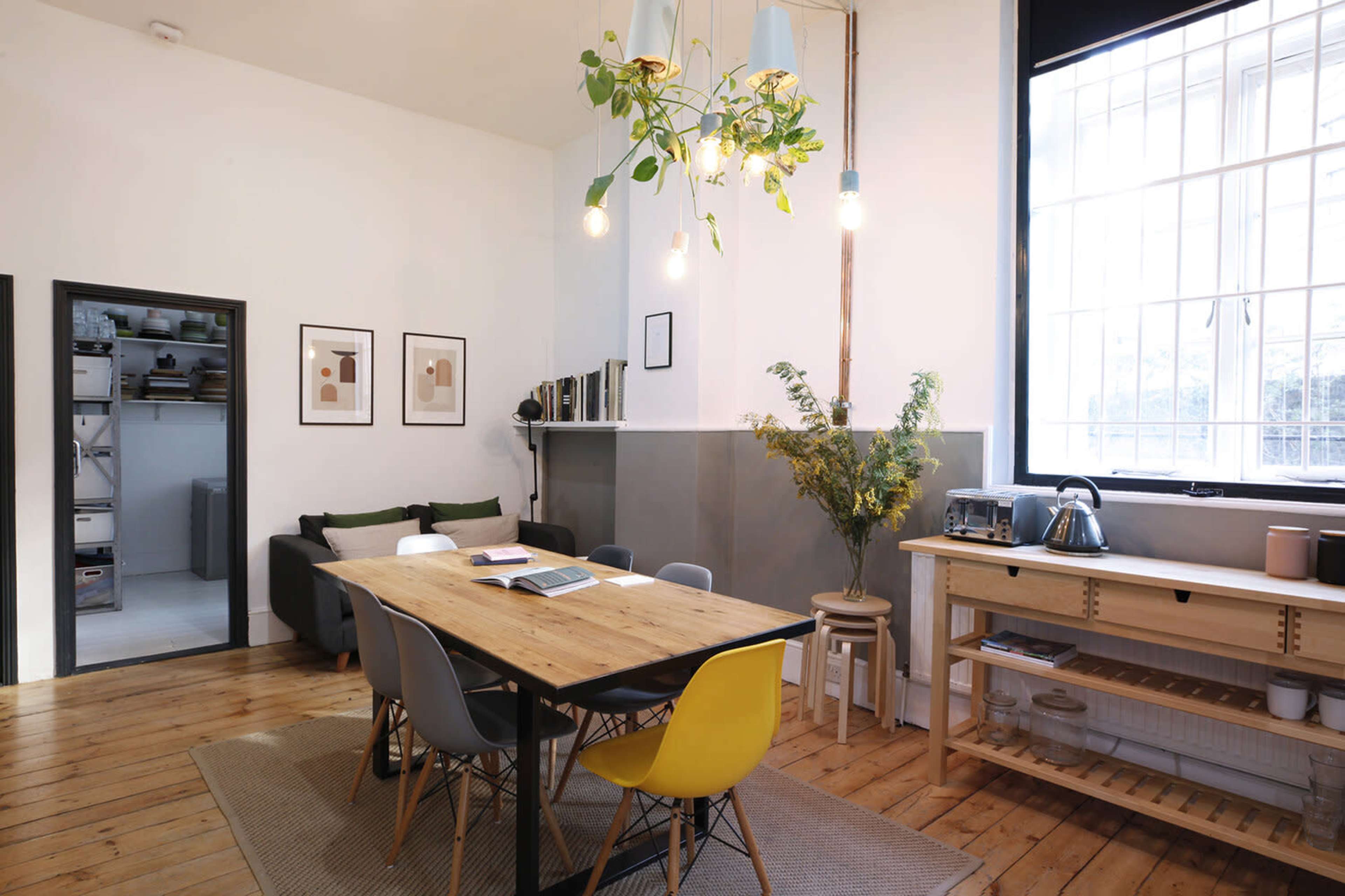 The image shows a modern dining area with a wooden table surrounded by colorful chairs, a plant hanging from the ceiling, and a large window providing natural light.