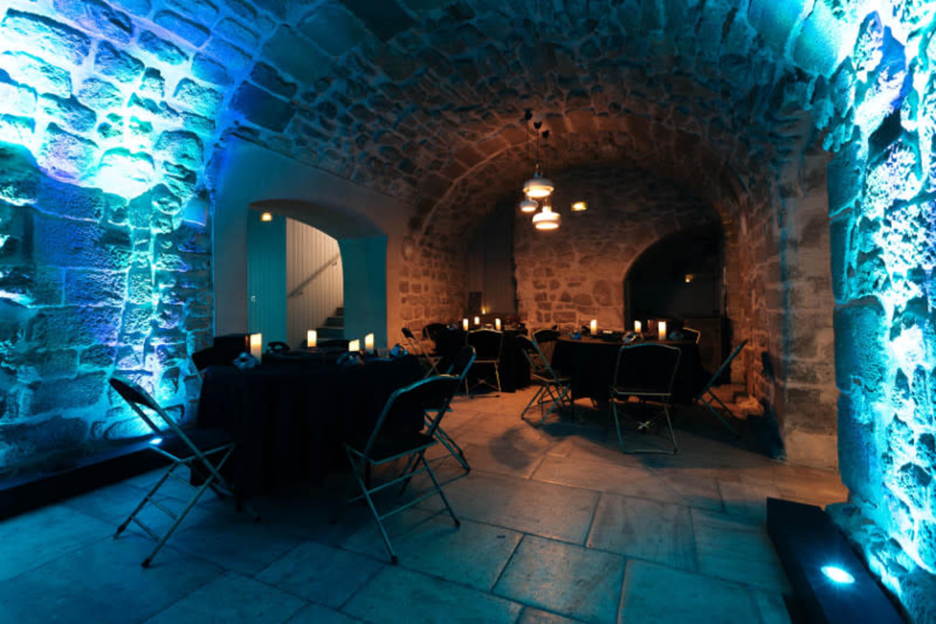 A dimly lit stone archway room with tables and chairs arranged for an event, illuminated by blue lighting.