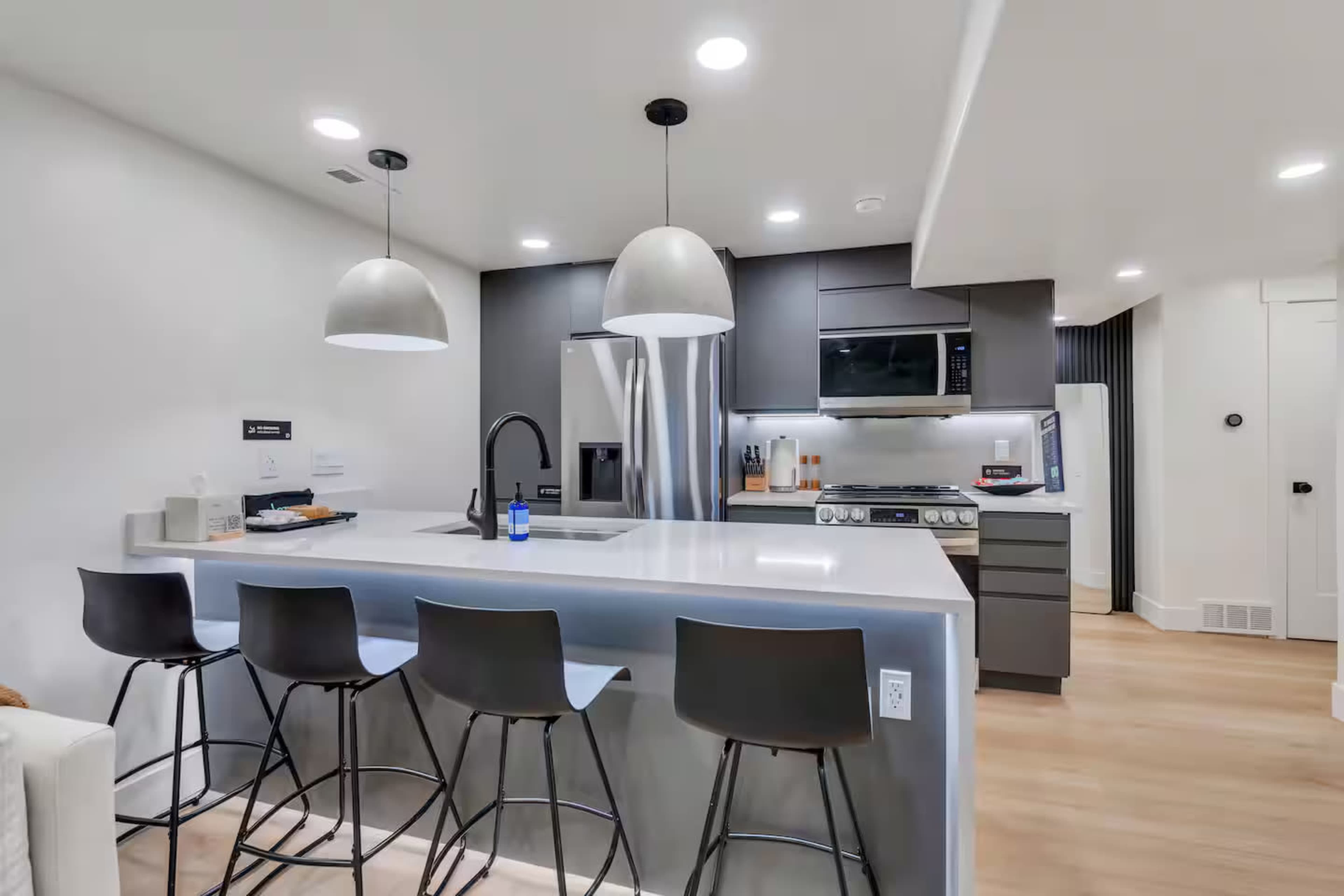 The image shows a modern kitchen featuring gray cabinetry, a stainless steel refrigerator, and a large island with four black bar stools.