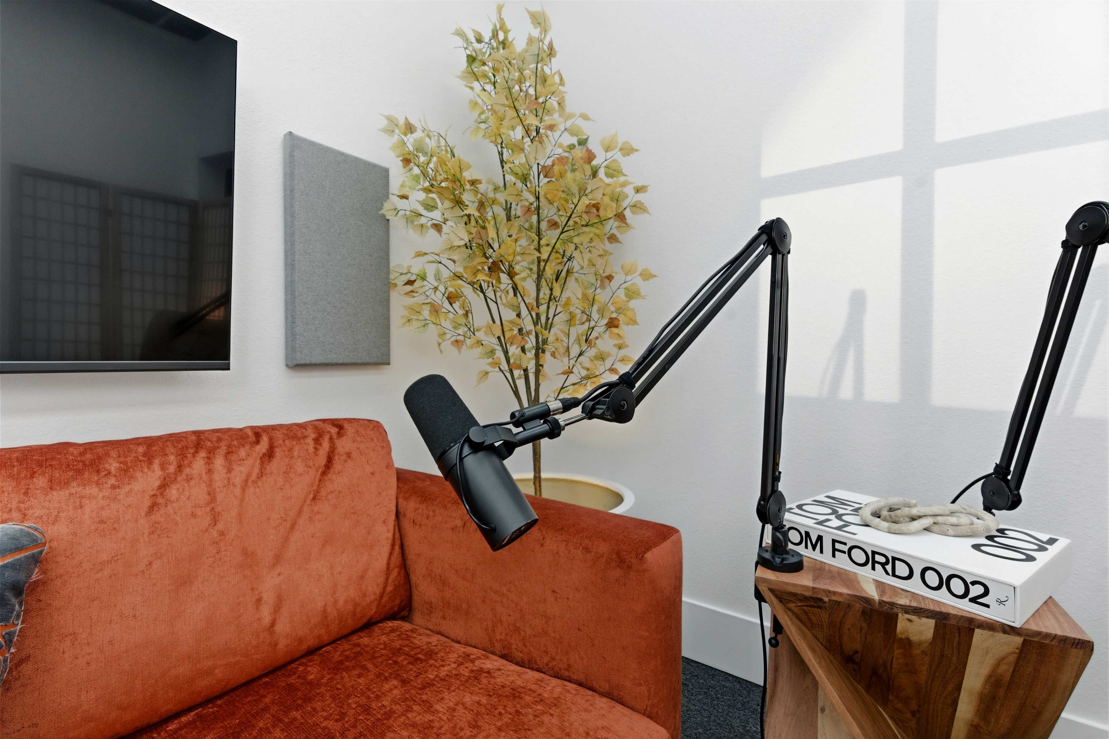 The image shows a cozy corner featuring an orange couch, a microphone on an adjustable arm, a decorative plant, and a stack of magazines on a small table.