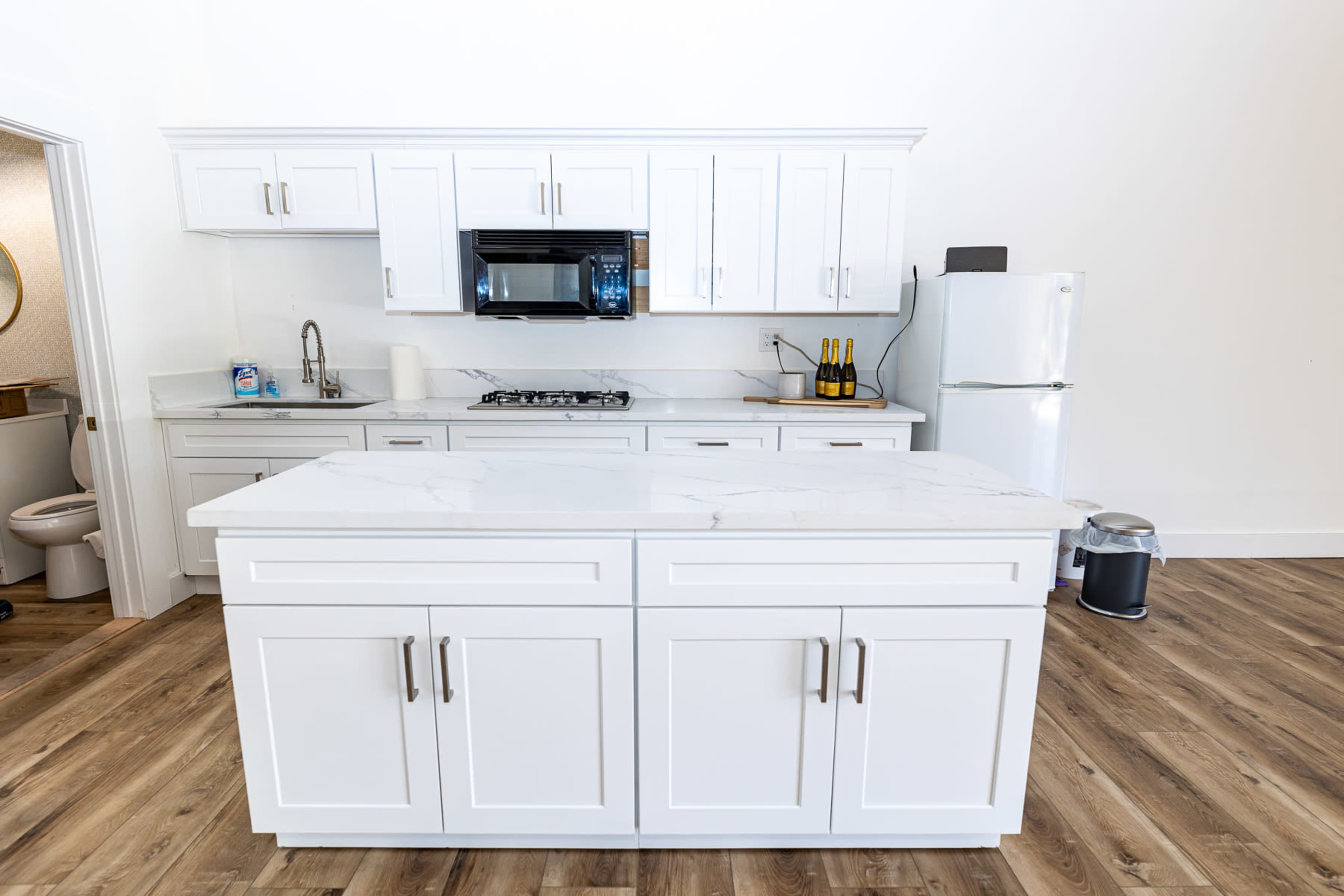 A modern kitchen features white cabinetry, a marble countertop island, stainless steel appliances, and wooden flooring.