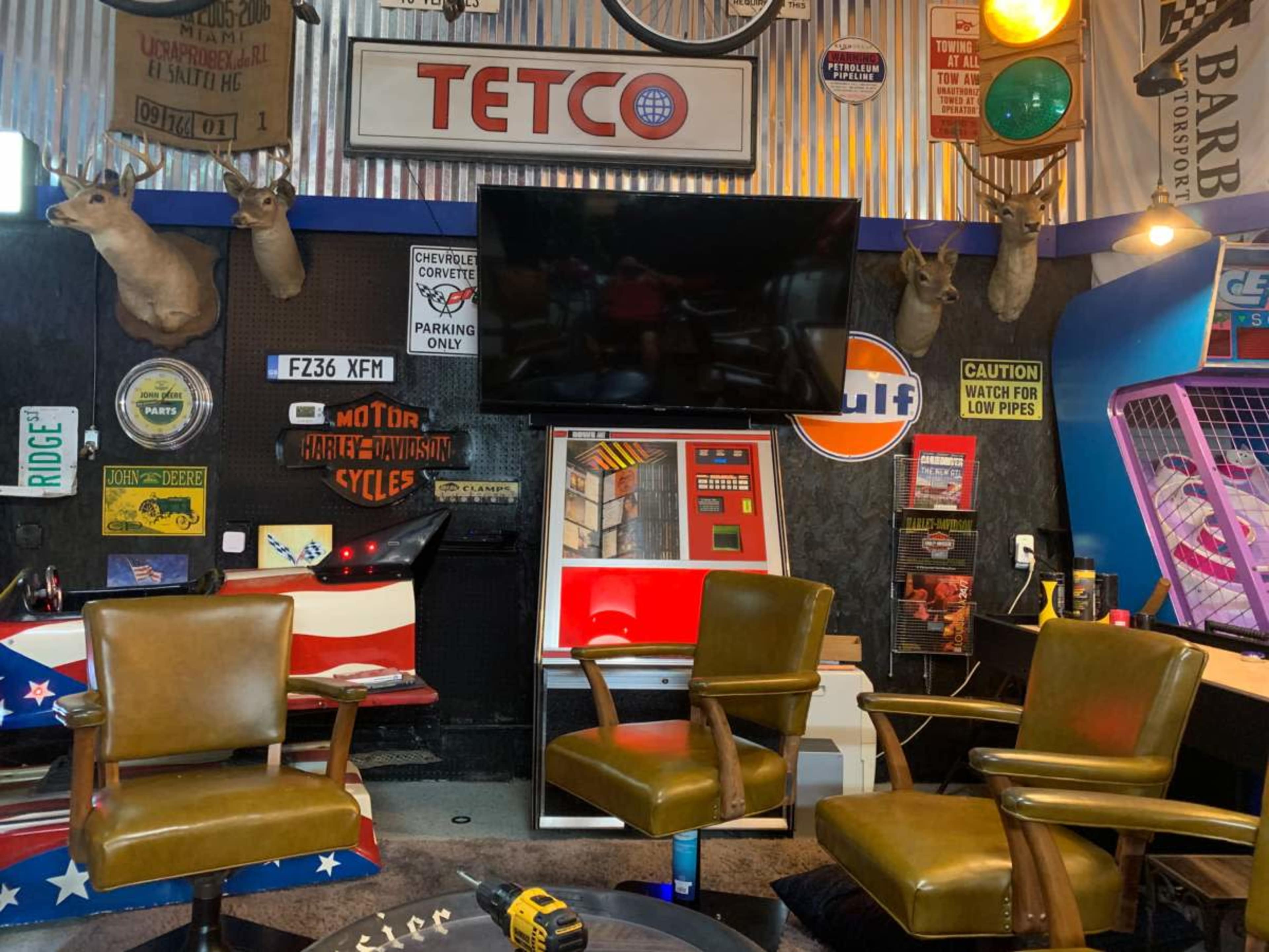 A rustic room decorated with vintage signs, taxidermy mounts, and various retro furnishings, including three brown leather chairs surrounding a central area with a television and arcade machine.