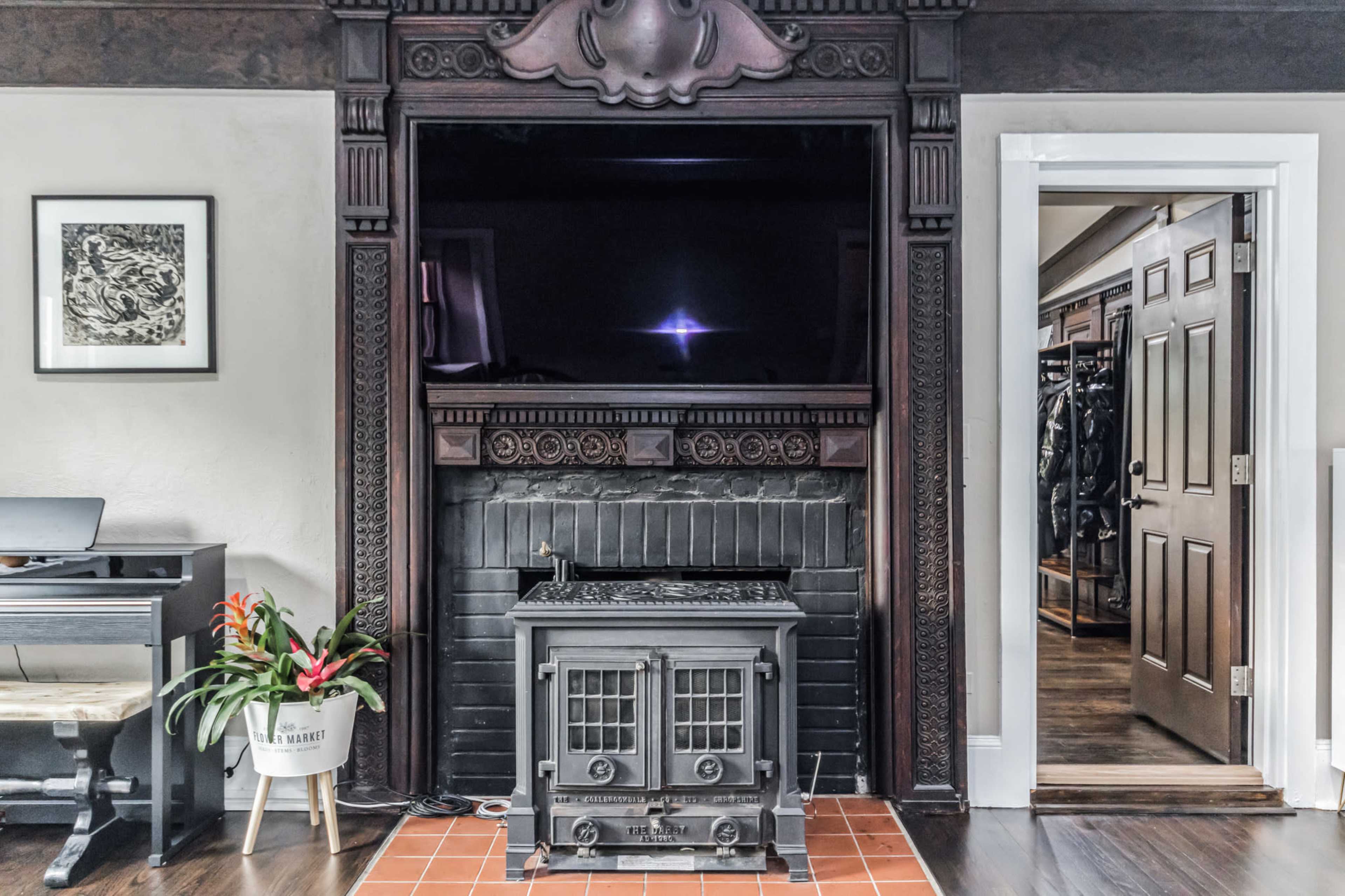 The image shows a room featuring an ornate fireplace mantle with a modern television mounted above it, a decorative plant in a white pot nearby, and a door leading to a closet on the right.