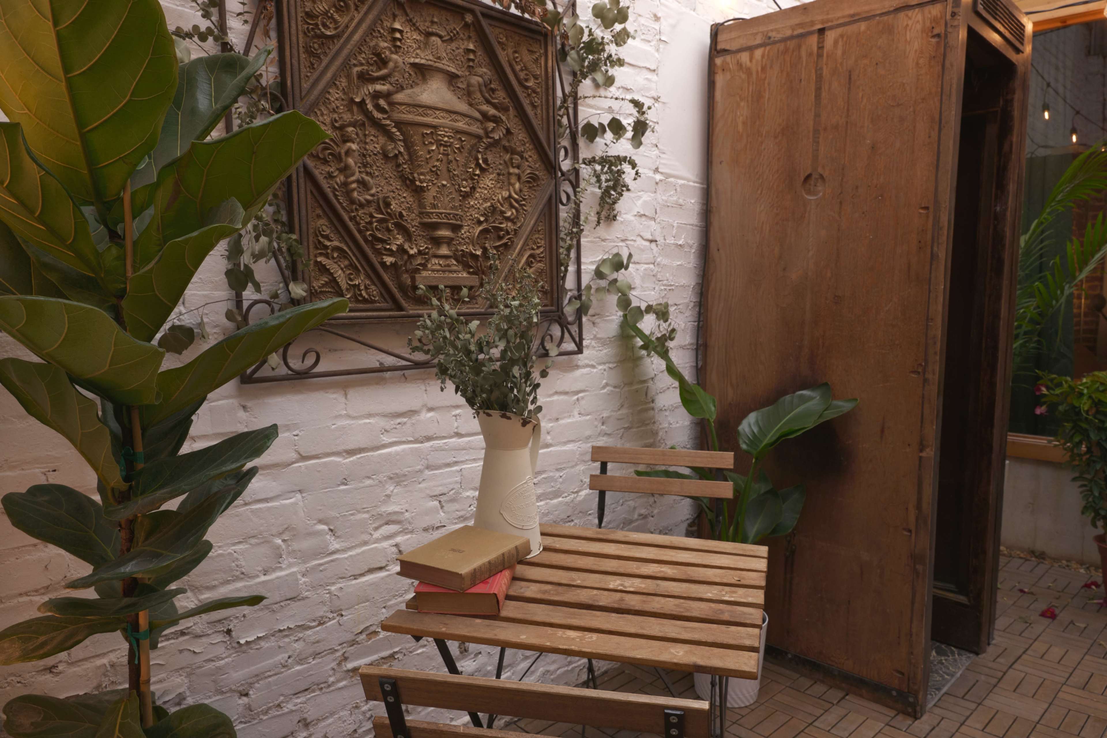 The image shows a small, cozy corner with a wooden table and chairs, a potted plant, a decorative wall piece, and an old wooden wardrobe.
