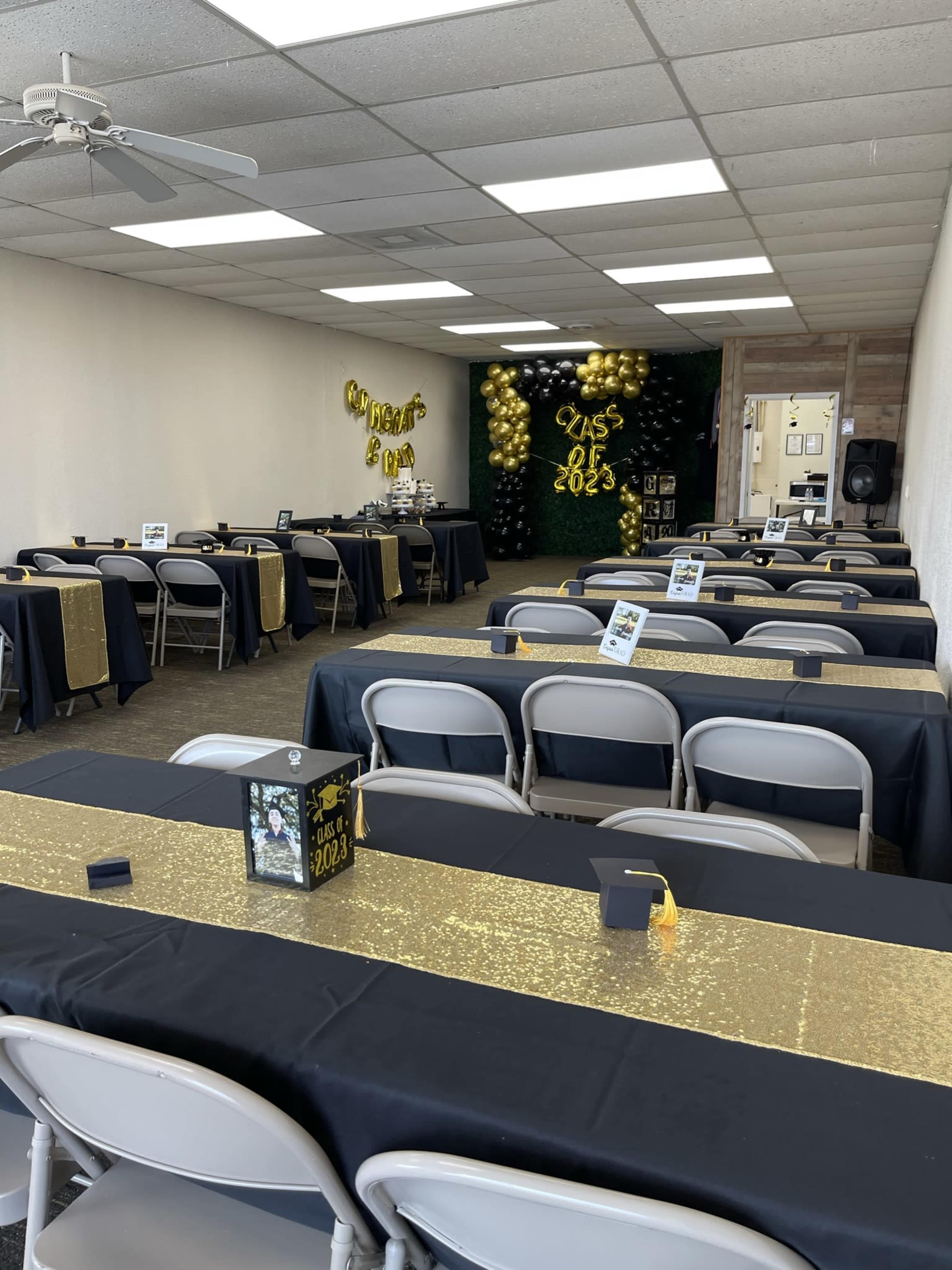 A decorated hall set up for a graduation celebration, featuring several tables with black and gold tablecloths, chairs, and balloons on the walls.
