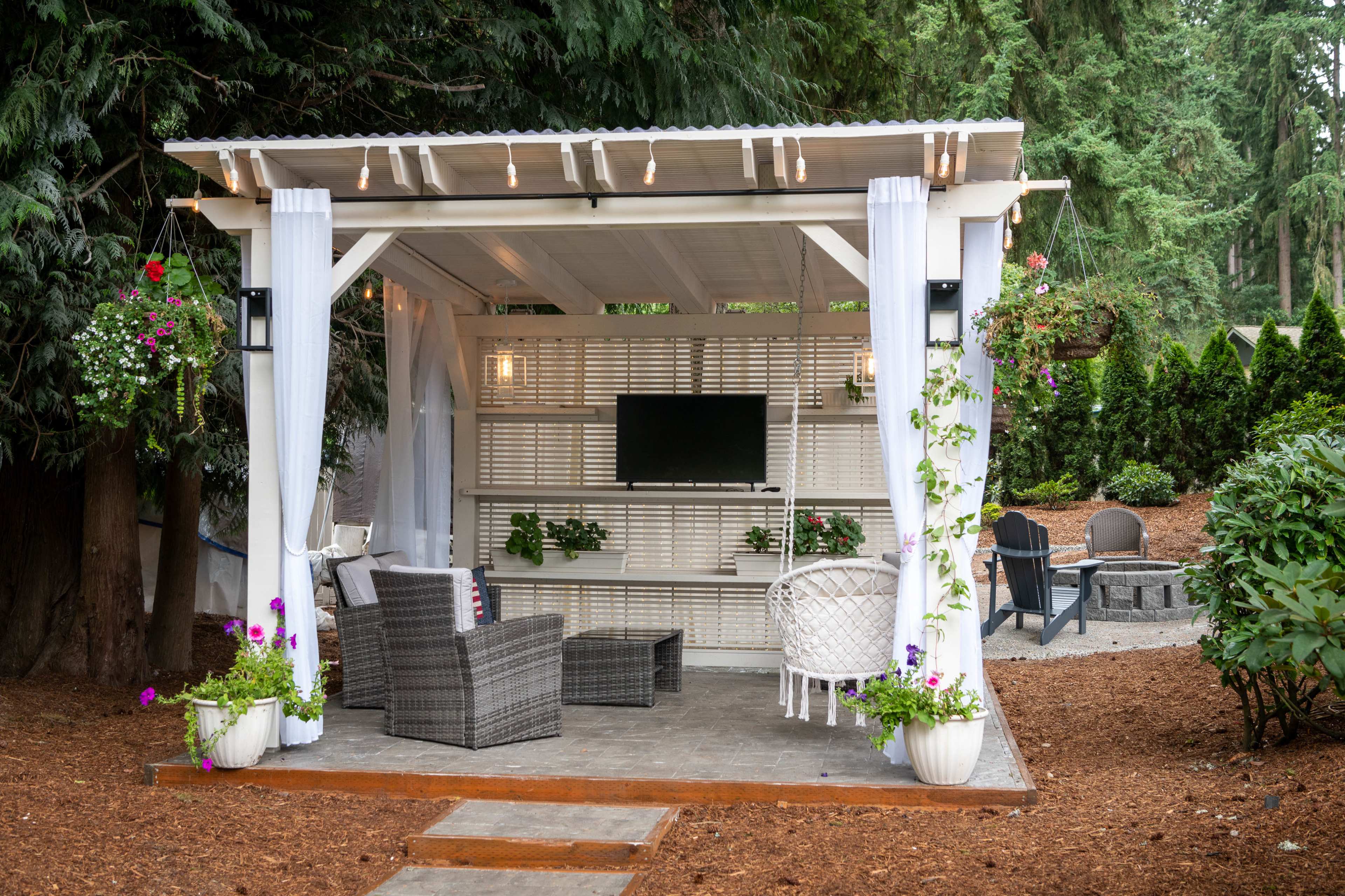 A cozy outdoor pavilion features comfortable seating, a wall-mounted television, and decorative plants, set against a backdrop of trees.