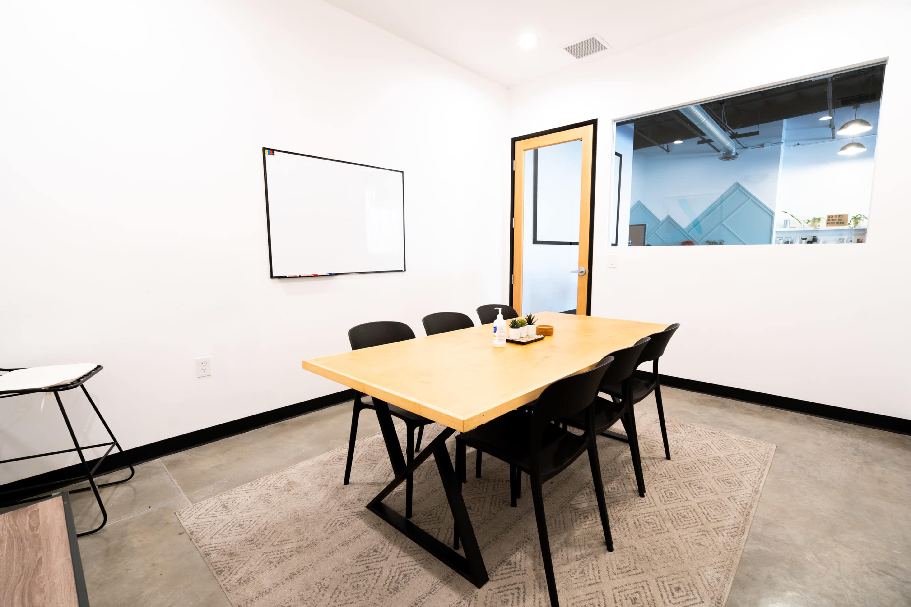 A meeting room with a rectangular wooden table and six black chairs, a whiteboard on one wall, and a glass door leading to another space.