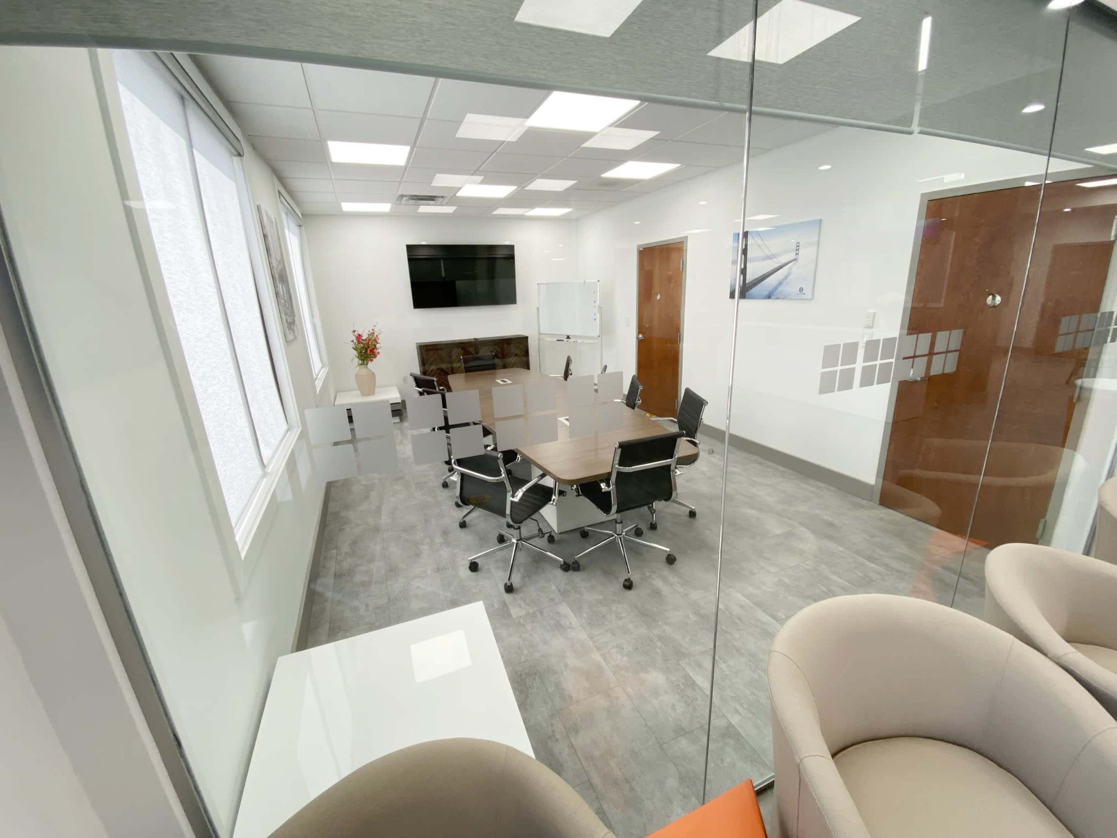 A modern conference room features a large table surrounded by rolling chairs, a TV screen on the wall, and glass partitions.