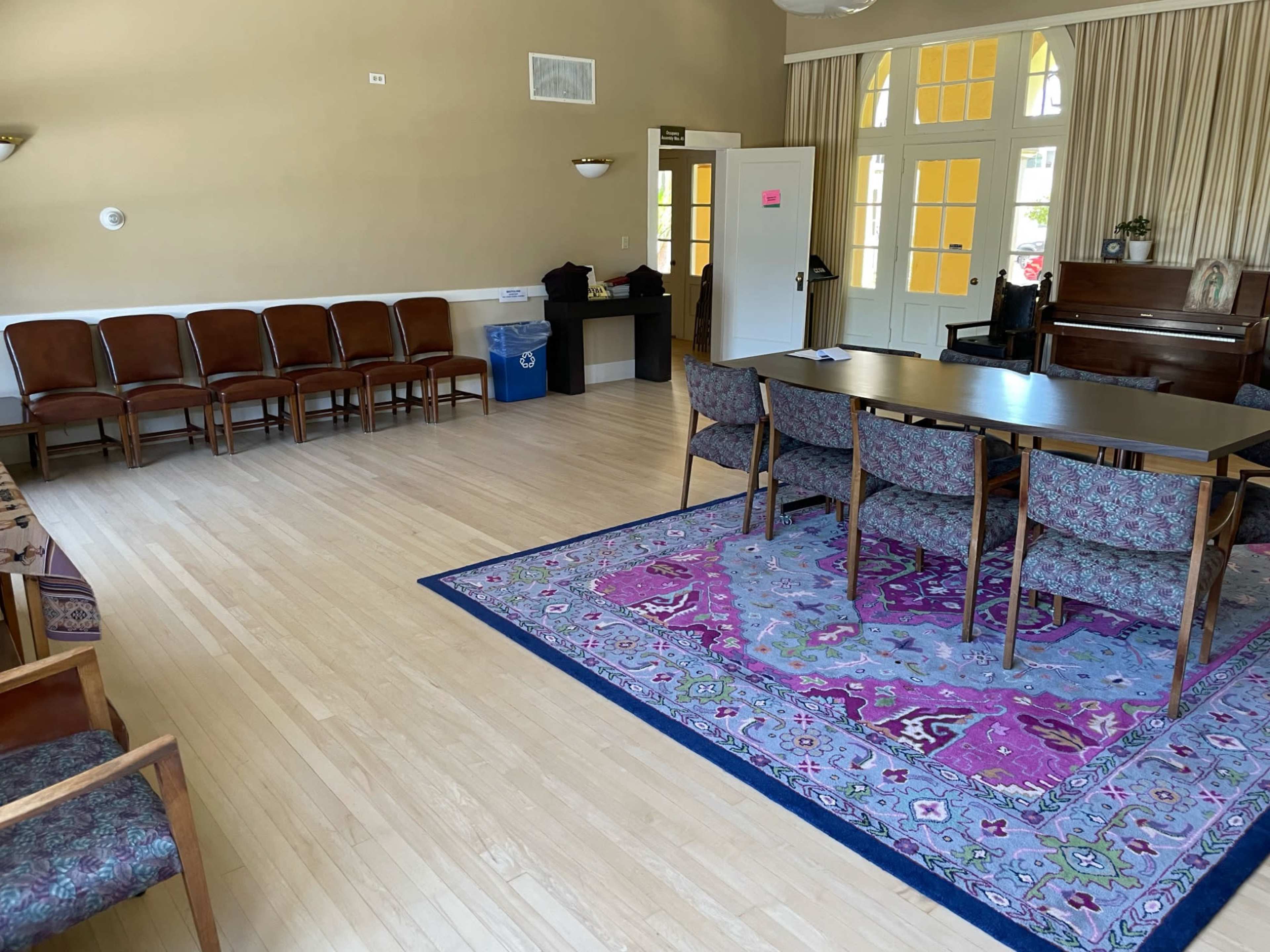 The image shows a spacious room with wooden flooring, a large table surrounded by chairs, benches along one wall, and a piano in the corner.